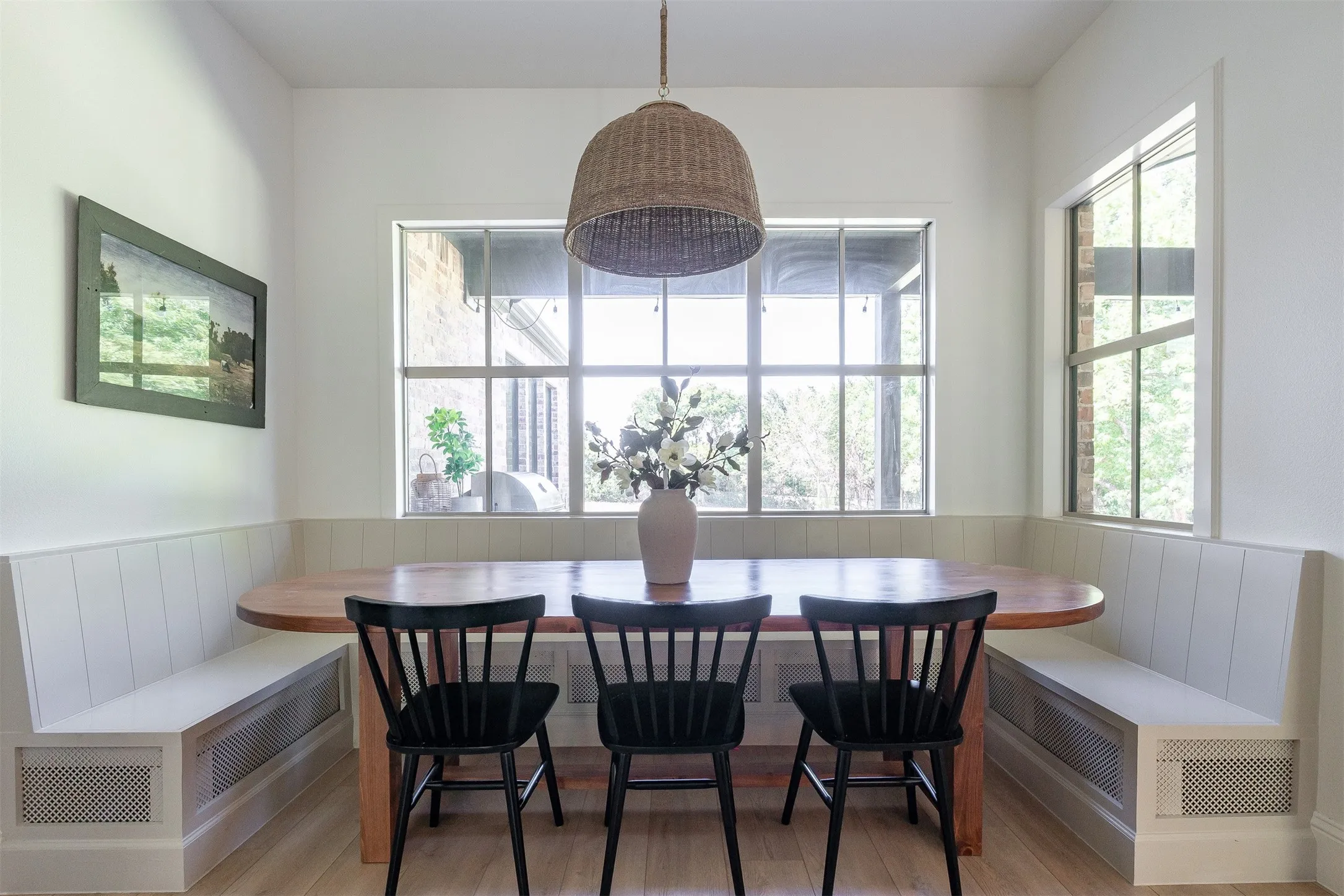 Built-in banquette seating with integrated grilles, white wainscoting, light-toned wood-finish flooring, and multiple windows with grids