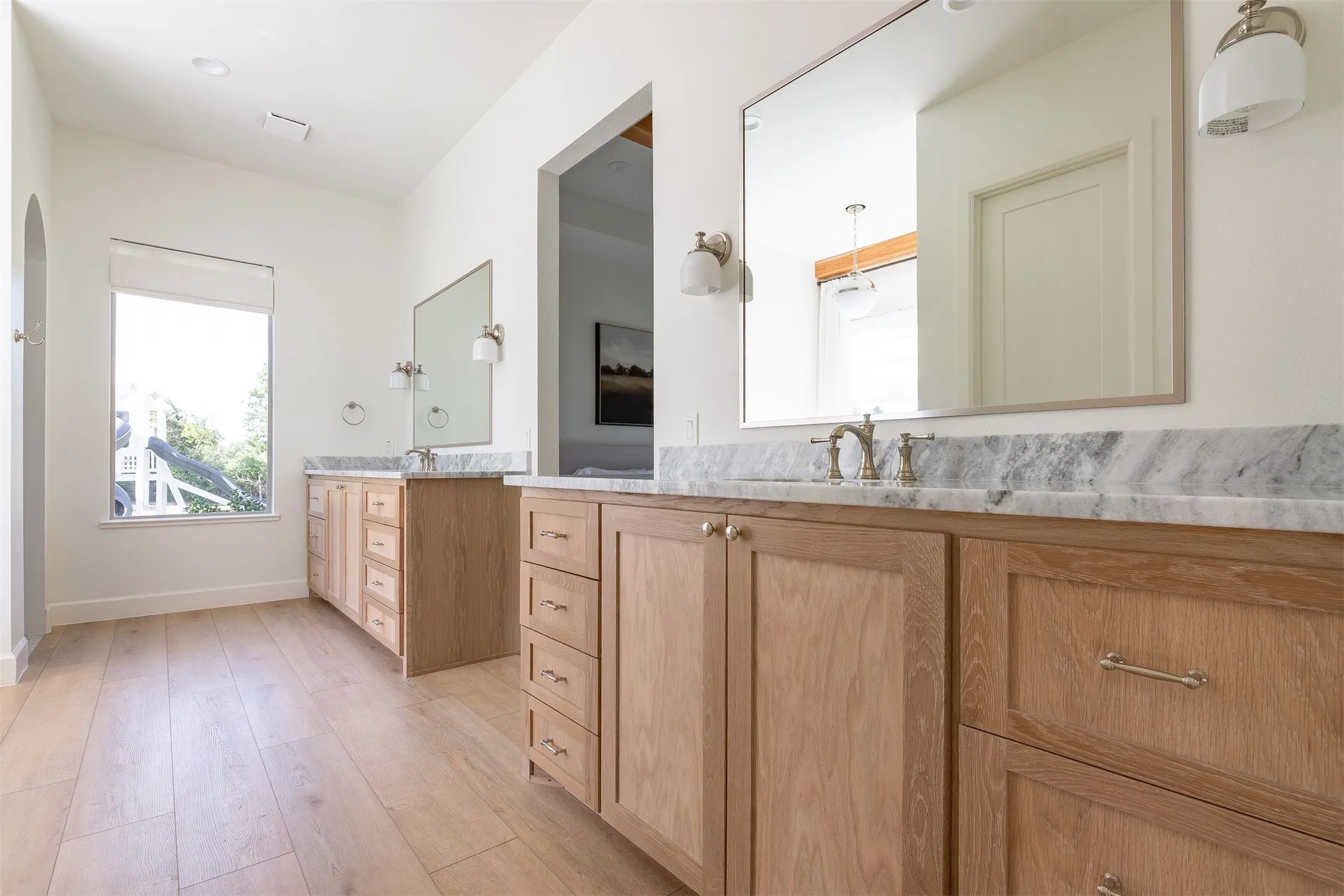 Dual wood-finish vanities featuring a light-toned wood finish, stone countertops, and integrated sinks with wall-mounted mirrors and sconce lighting