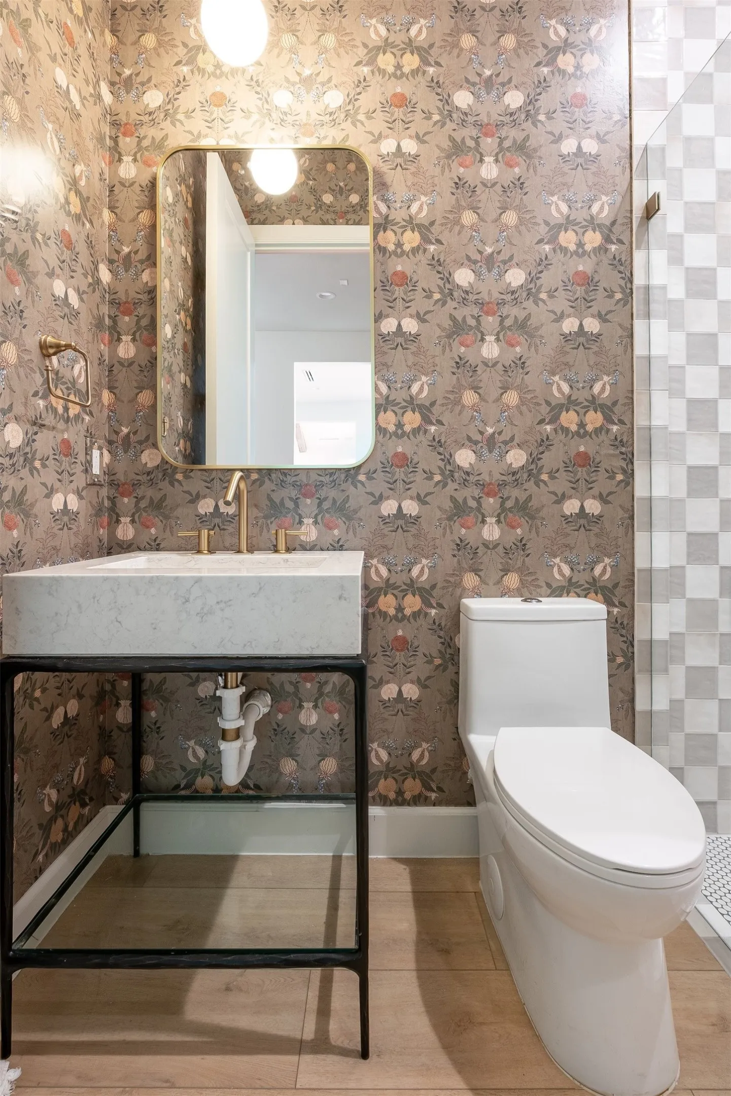 Full bath featuring botanical-patterned wallpaper, a gold-framed mirror, a vanity with a rectangular basin, and wood-finish flooring