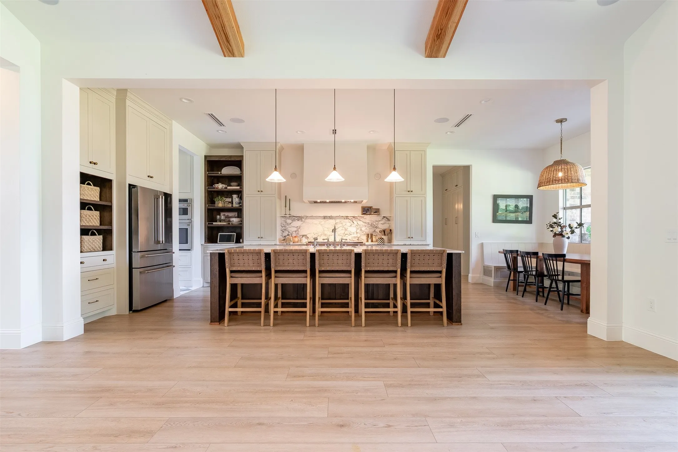 Open-concept kitchen with a large central island, light wood-finish flooring, exposed ceiling beams, built-in cabinetry, and stainless steel appliances
