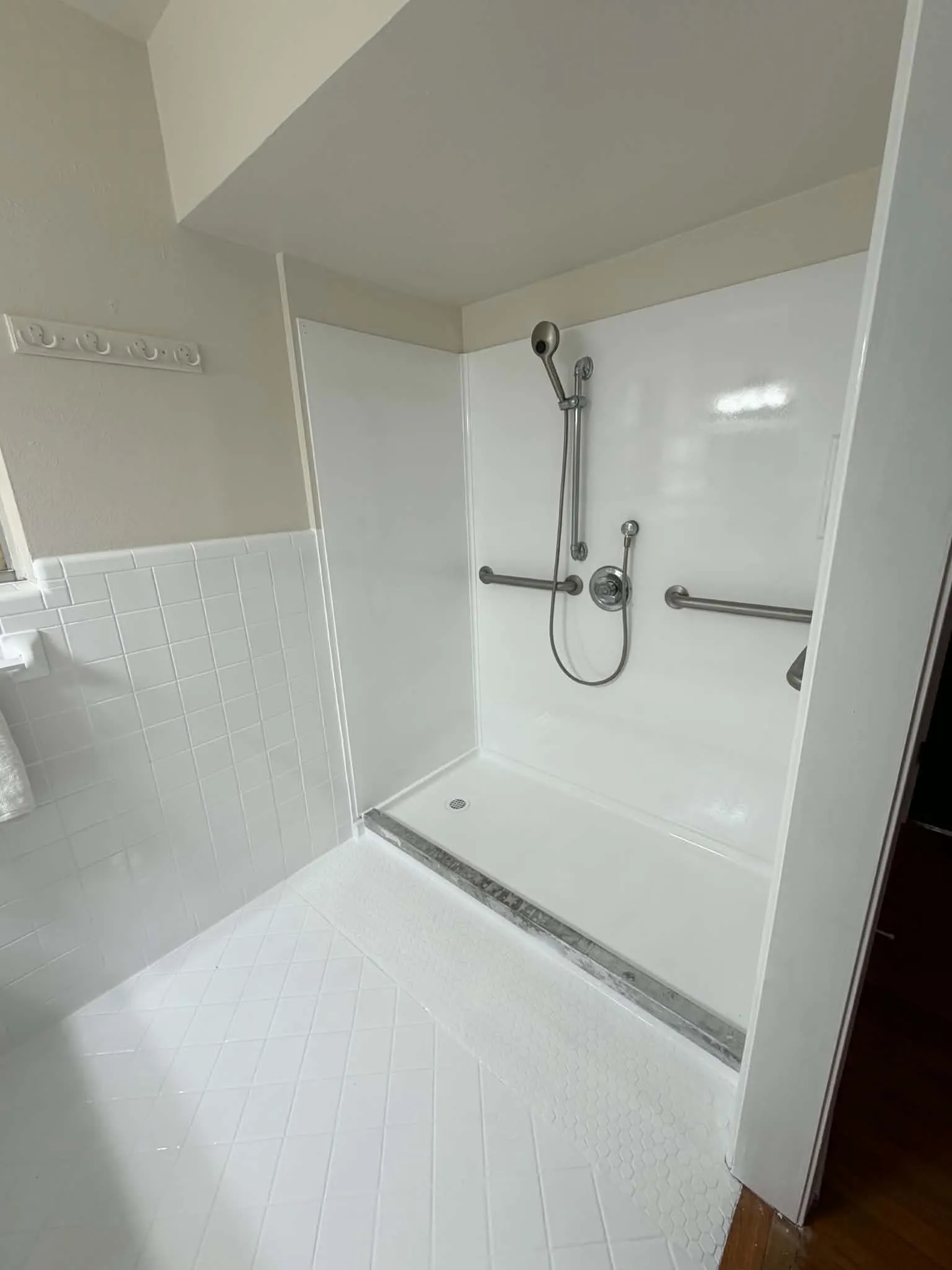 Shower with white paneling and a flexible shower head, featuring a textured tile floor and grab bars