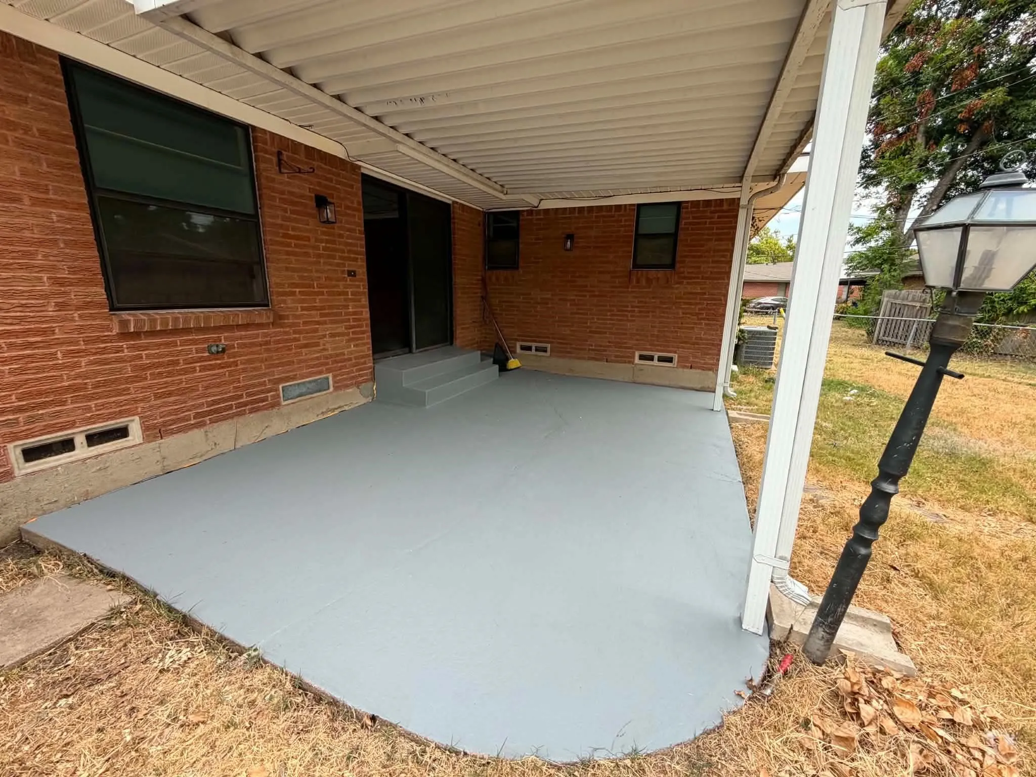 Covered concrete patio with a brick exterior, featuring a sliding glass door and a wall-mounted light fixture