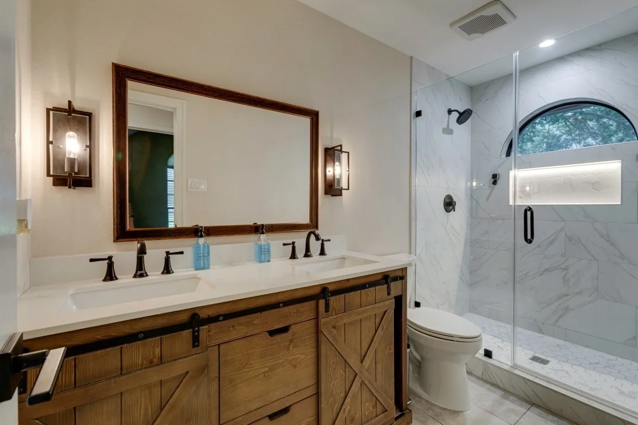 Dual vanity with a white countertop and matte black fixtures, barn door-style cabinet, walk-in shower with glass enclosure, arched window, and marble-look wall tile