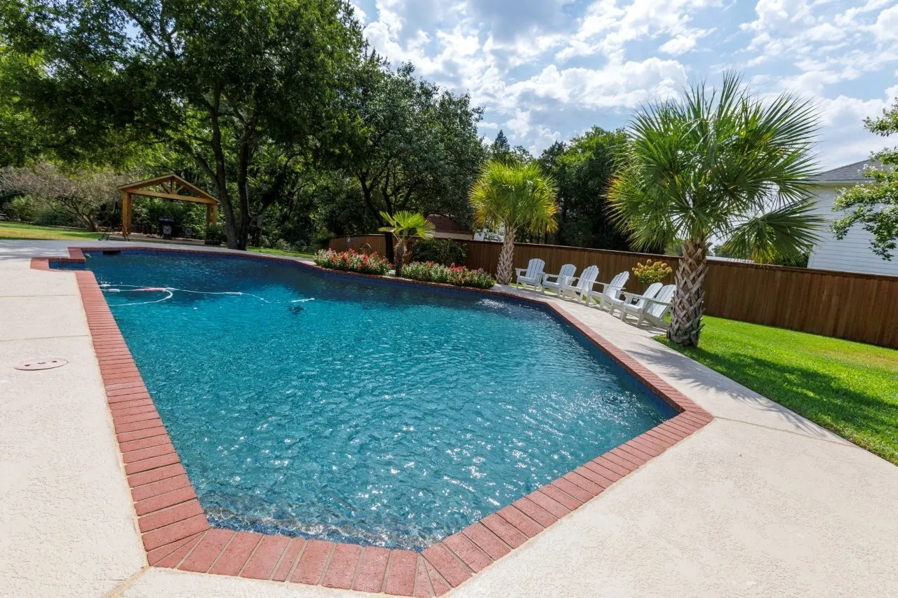 Swimming pool with a brick coping, an expansive concrete deck, mature palm trees, and a wooden privacy fence