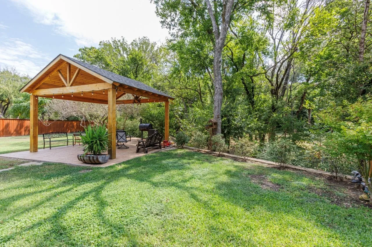 Expansive green lawn featuring a wood-framed outdoor pavilion with a shingled roof, string lighting, and a paved patio area