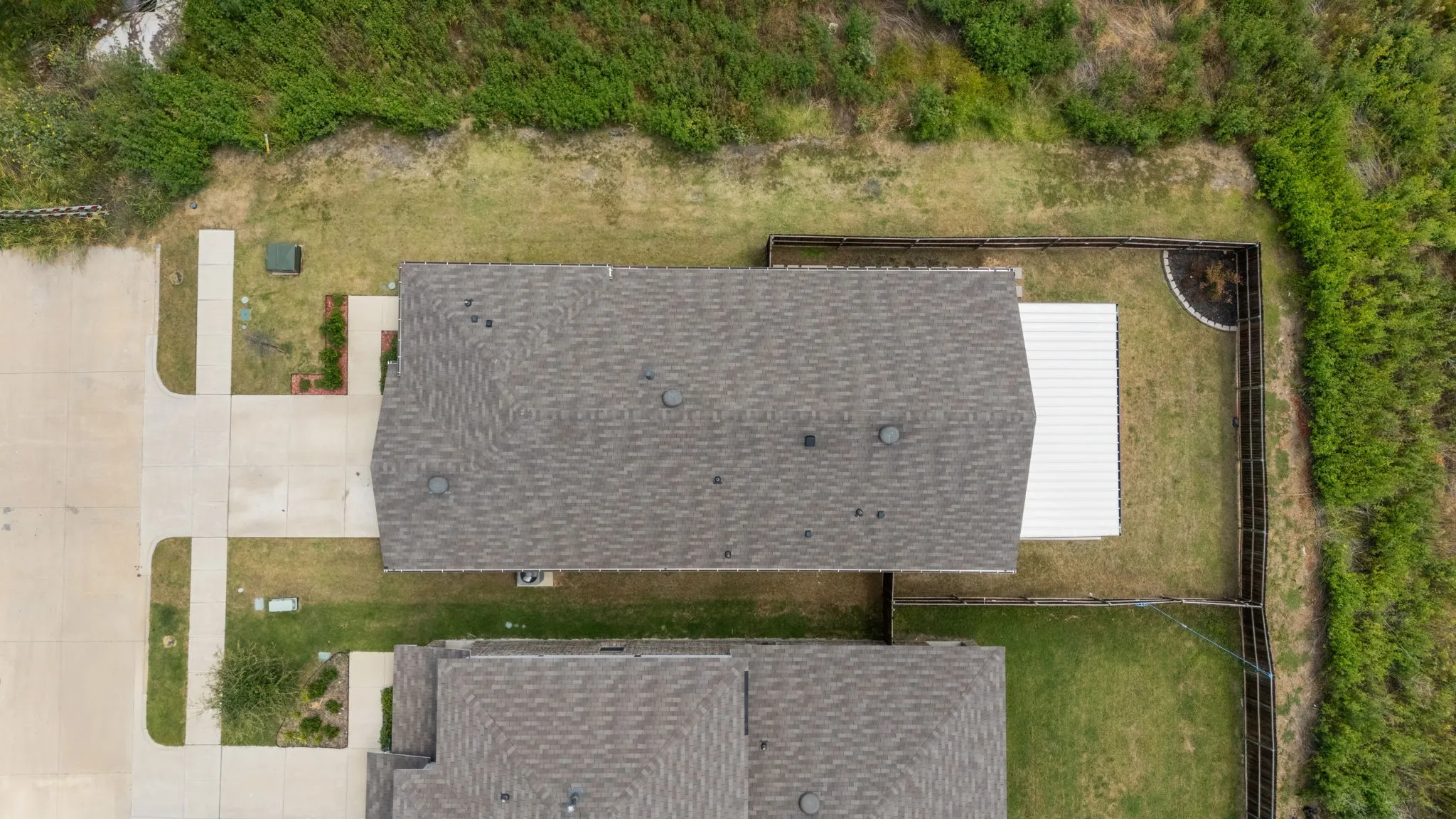 Gabled roofline with asphalt shingles, concrete driveway, concrete walkways, wood fencing, and a white metal roof structure
