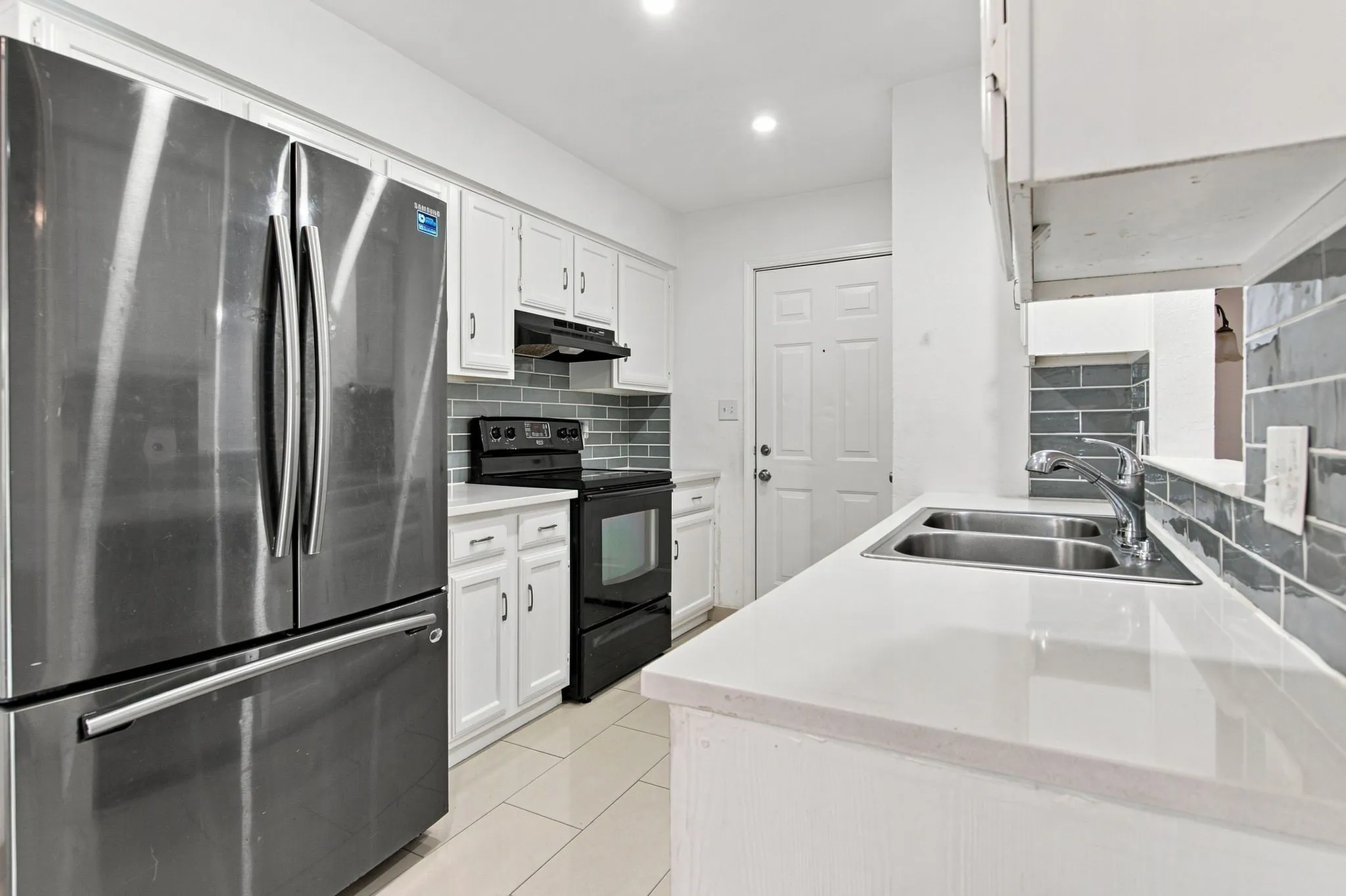 Stainless steel French door refrigerator, white cabinetry, solid surface countertops, subway tile backsplash, and a double basin sink