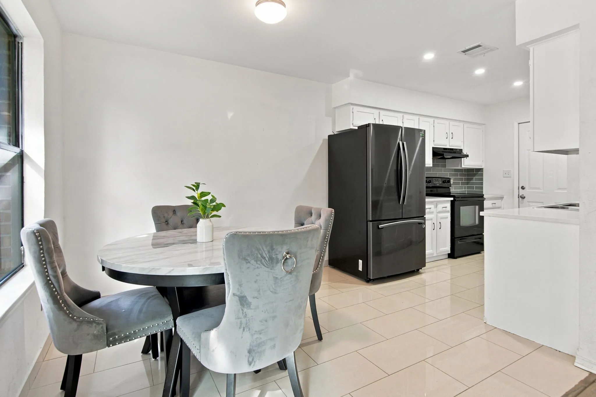 Bright dining area with tile flooring and a kitchen featuring white cabinetry, black appliances, and a gray subway tile backsplash