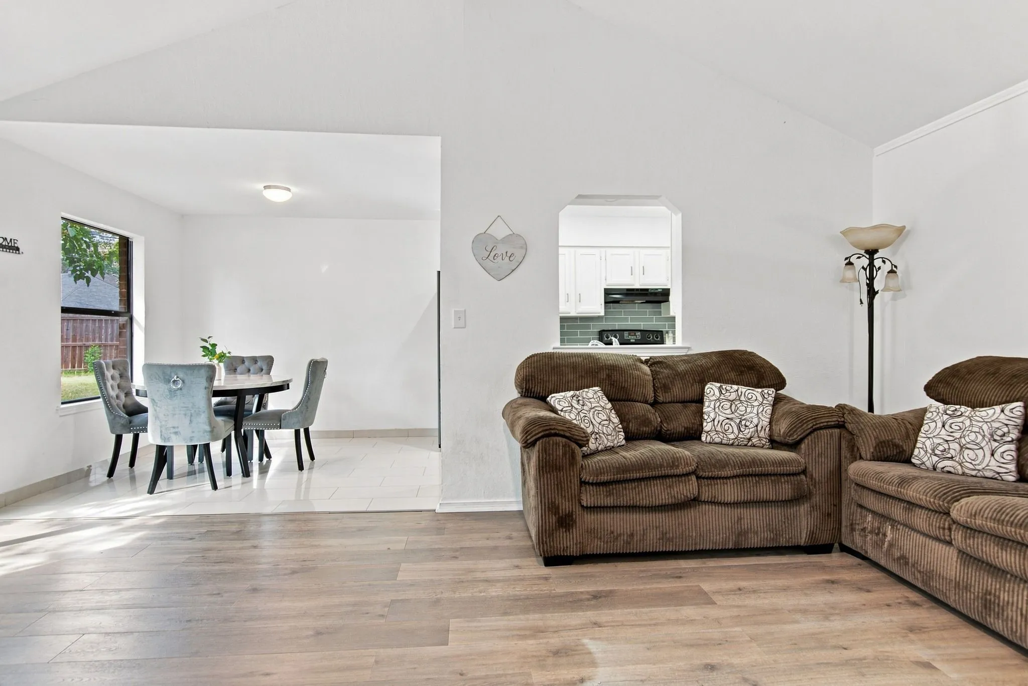 Vaulted ceiling, wood-finish flooring, white cabinetry, teal subway tile backsplash, and a pass-through window