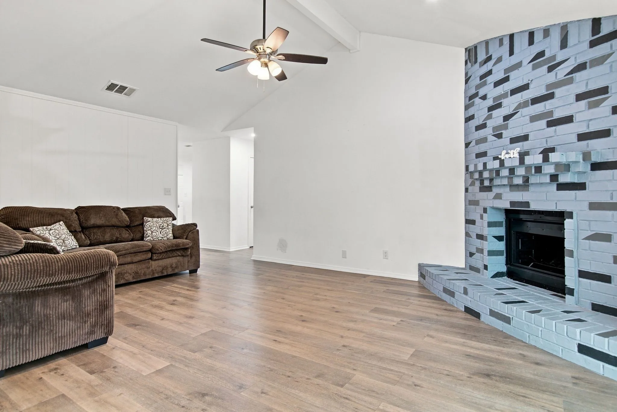 Vaulted ceiling with exposed beam, ceiling fan, wood-finish flooring, and a brick fireplace