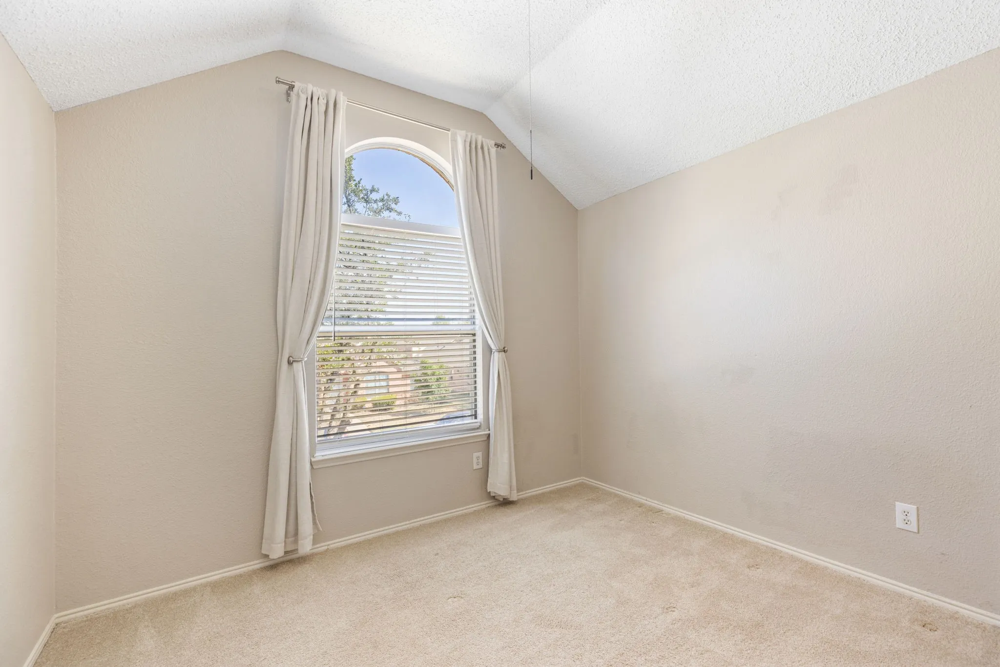 Vaulted ceiling room featuring an arched window with blinds, neutral carpeting, and textured walls