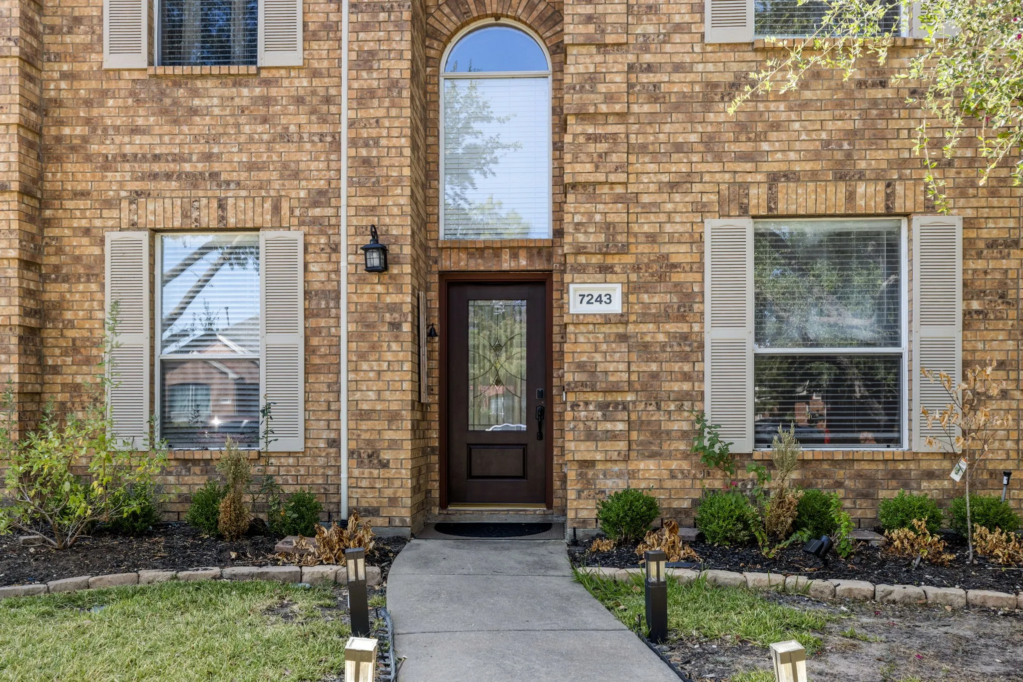 Brick exterior with decorative shutters, arched transom window, front entry door with glass insert, concrete walkway, and landscaped garden beds