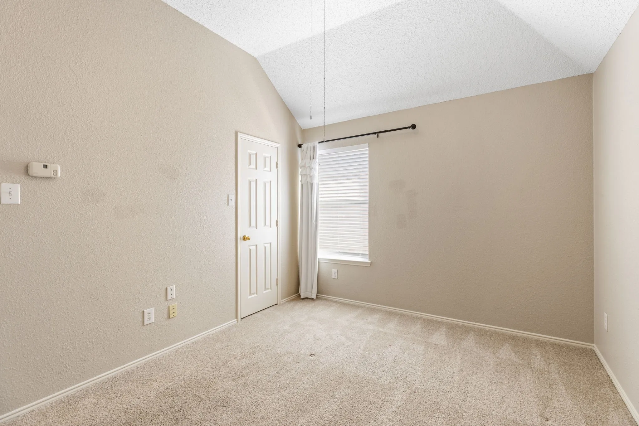 Vaulted ceiling with textured finish, carpeted flooring, window with horizontal blinds, paneled interior door, and neutral-toned walls