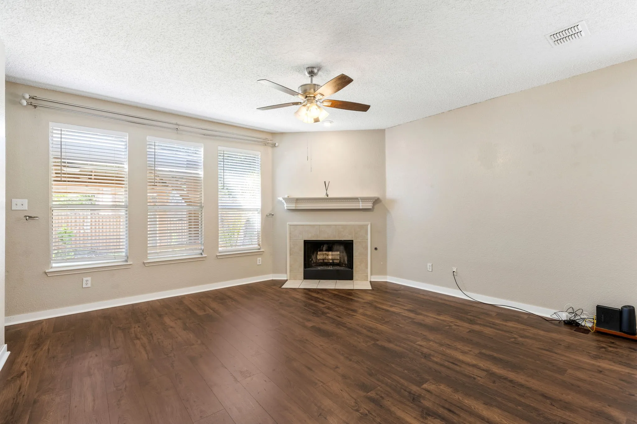 Wood-finish flooring, a tiled fireplace with a mantel, multiple windows with blinds, and a ceiling fan