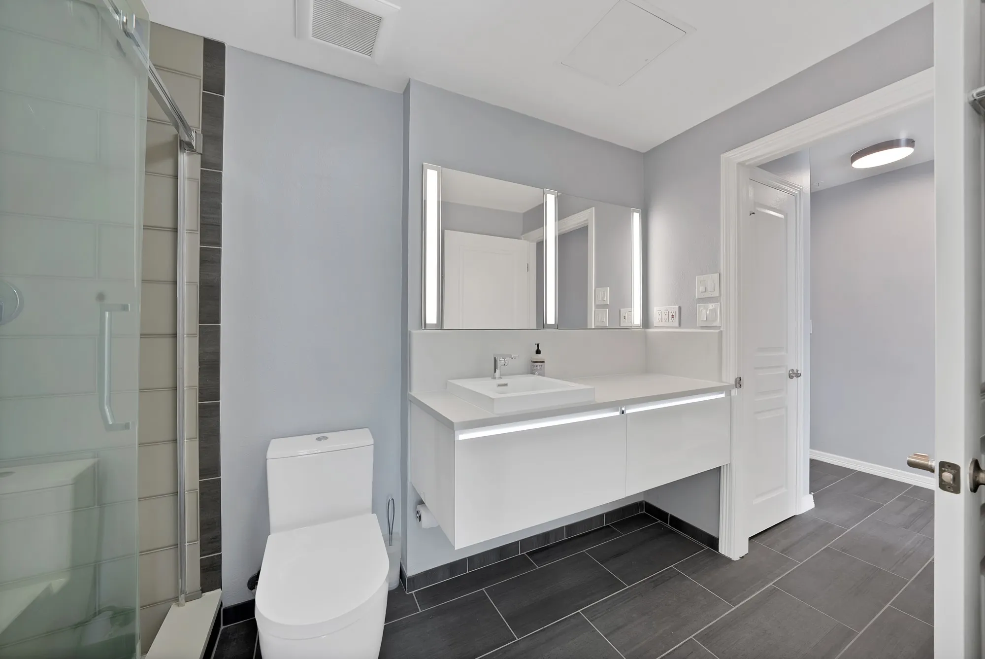 Glass-enclosed shower with tiled surround, floating vanity with integrated lighting, rectangular vessel sink, illuminated mirrored medicine cabinets, and dark-toned floor tiling