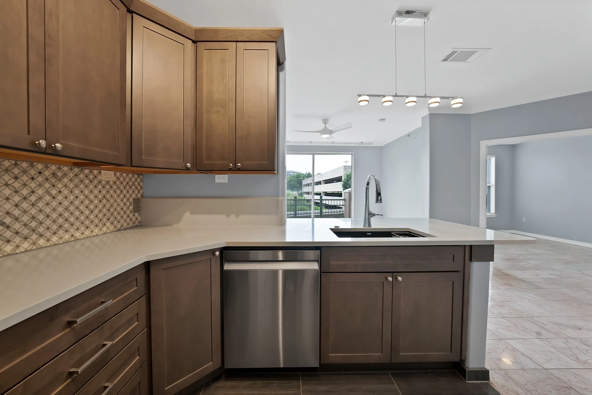 Full kitchen with wood cabinetry, stainless steel dishwasher, an integrated sink with a gooseneck faucet, light-toned countertops, and a patterned tile​​‌​​​​‌​​‌‌​‌‌​​​‌‌​‌​‌​‌​​​‌​​ backsplash