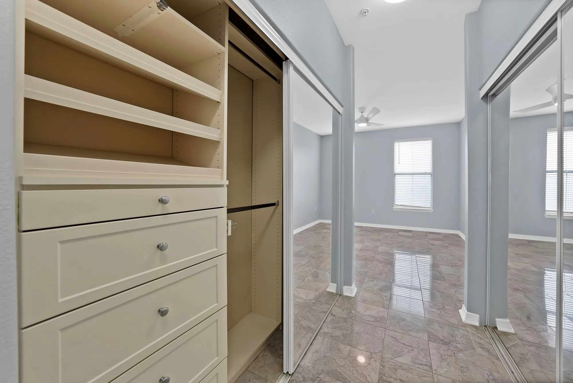 Built-in closet organization system with shelving and drawers, mirrored sliding closet doors, ceiling fan, and tile flooring