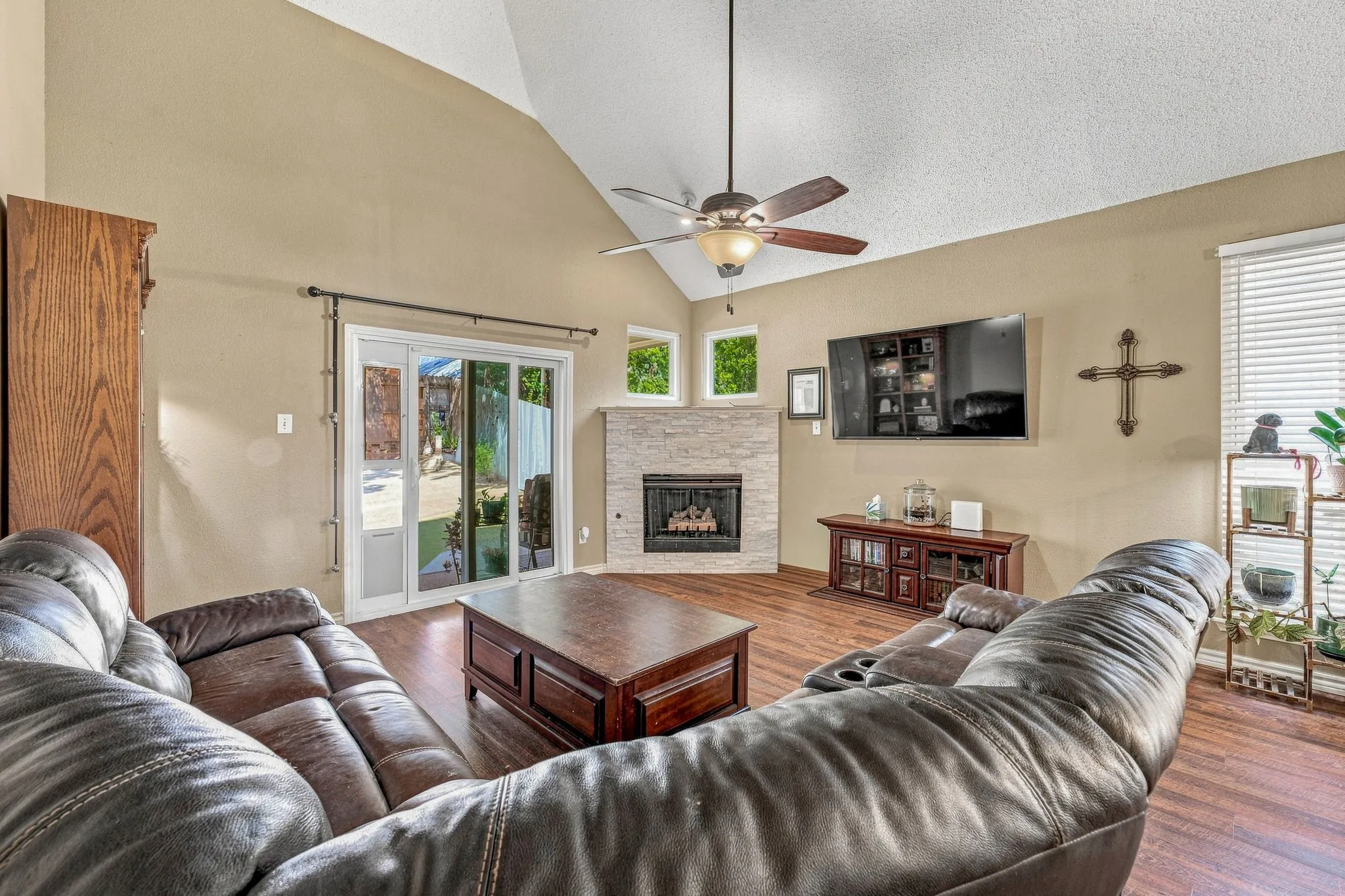 Vaulted ceiling, wood-finish flooring, stone-clad fireplace with hearth, ceiling fan, and sliding glass door