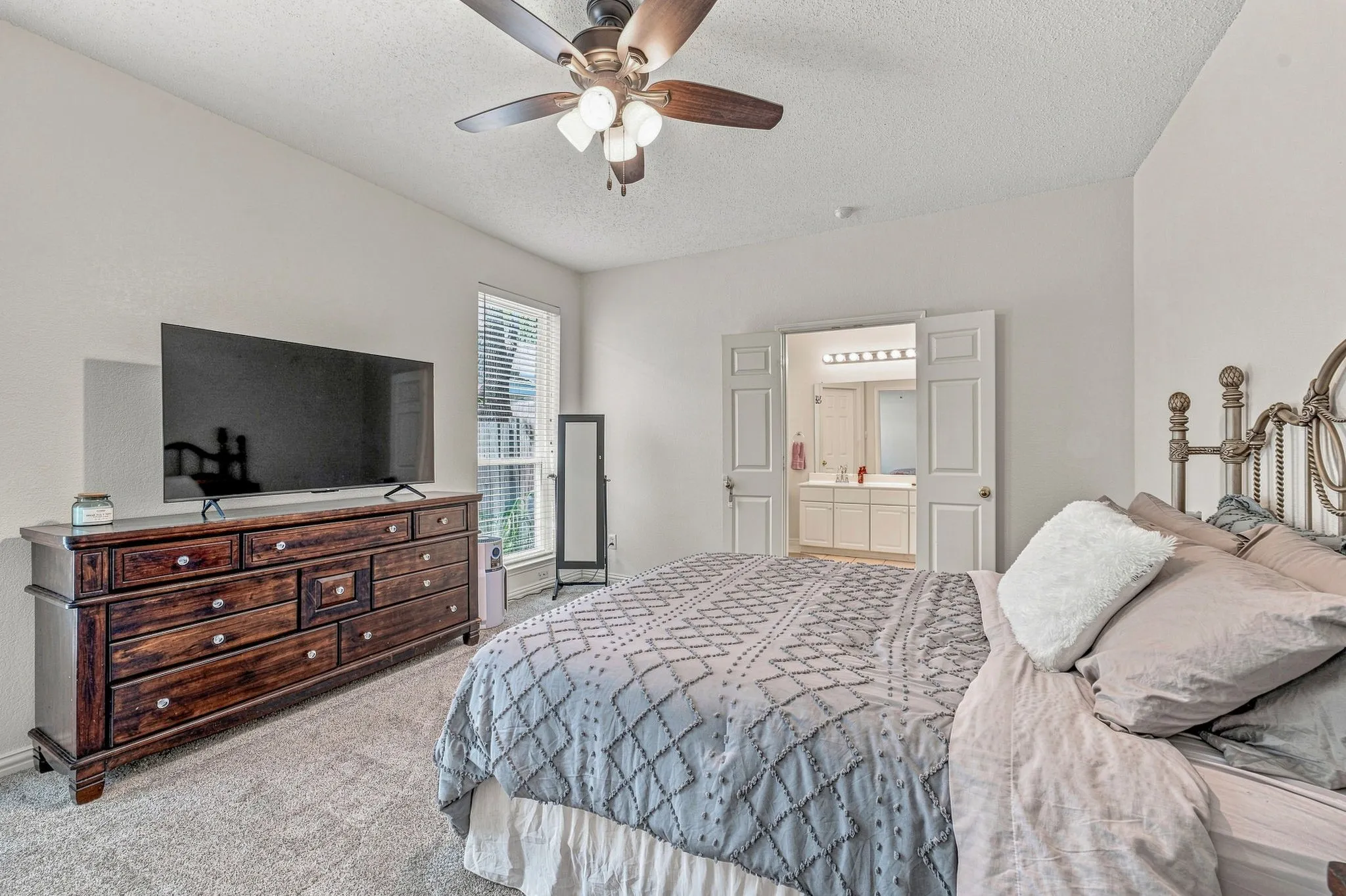 Neutral wall paint, carpeted flooring, window with blinds, ceiling fan with integrated lighting, and double-door entry to a bathroom