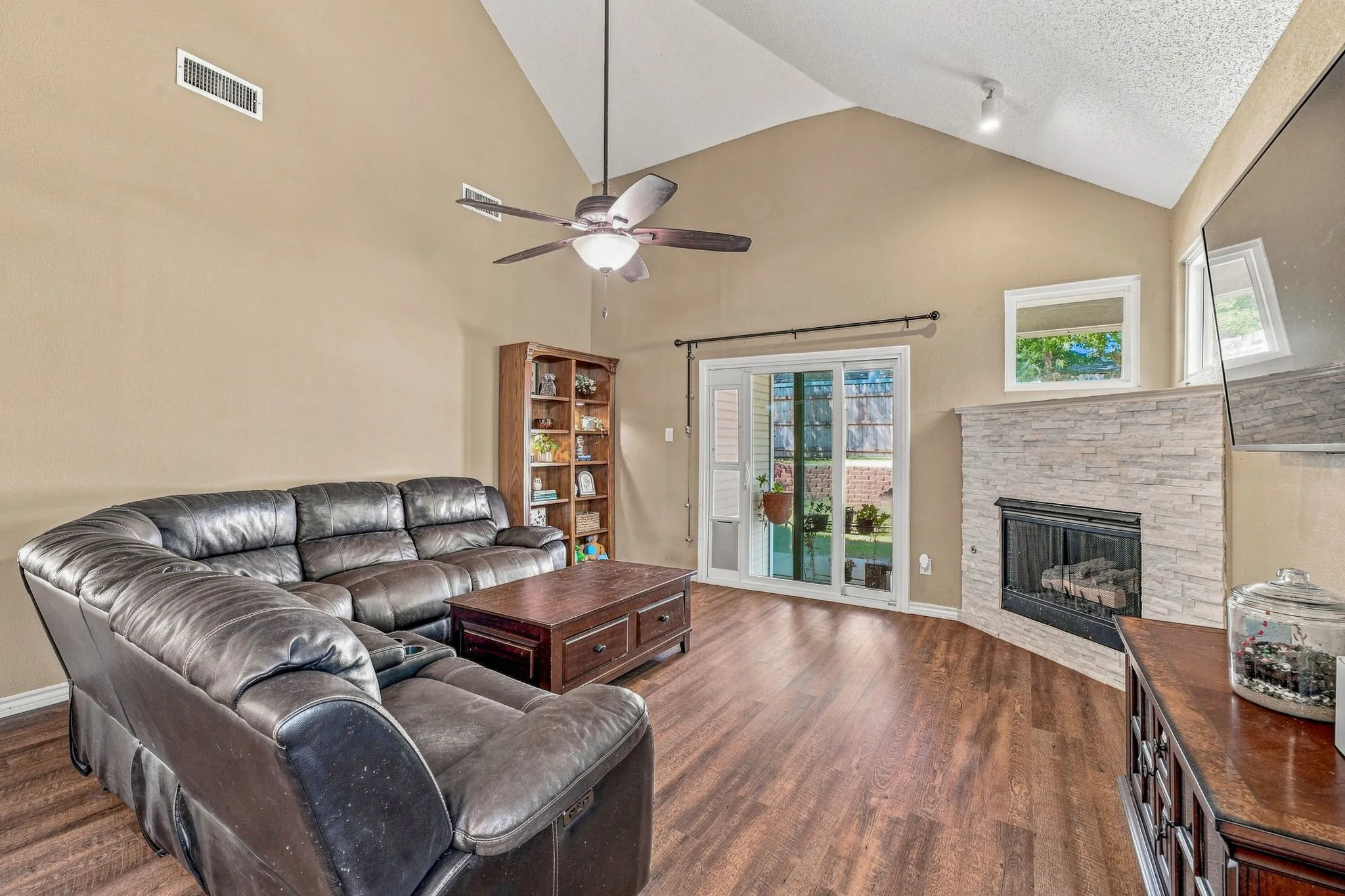 Vaulted ceiling with an installed ceiling fan, wood-finish flooring, a fireplace with a stacked stone surround, and sliding glass doors