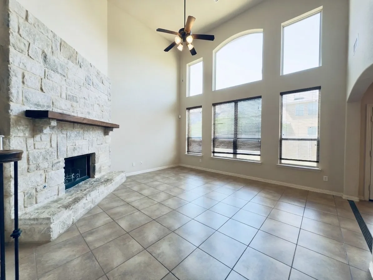 Stone-faced fireplace with a wood mantel, high vaulted ceilings, large arched window, multiple windows with blinds, and tile flooring