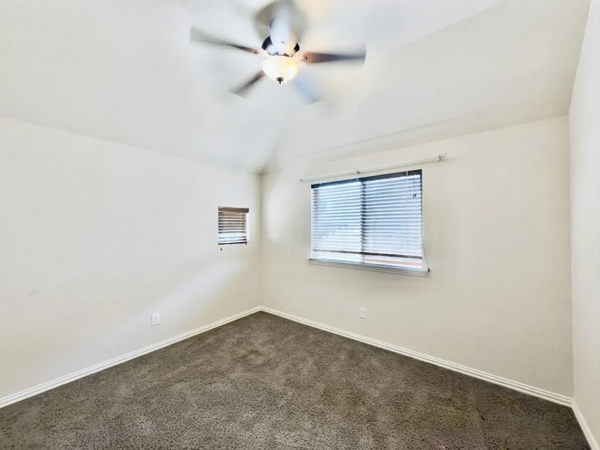 Vaulted ceiling with a flush-mount ceiling fan, two windows with blinds, and dark gray carpeting