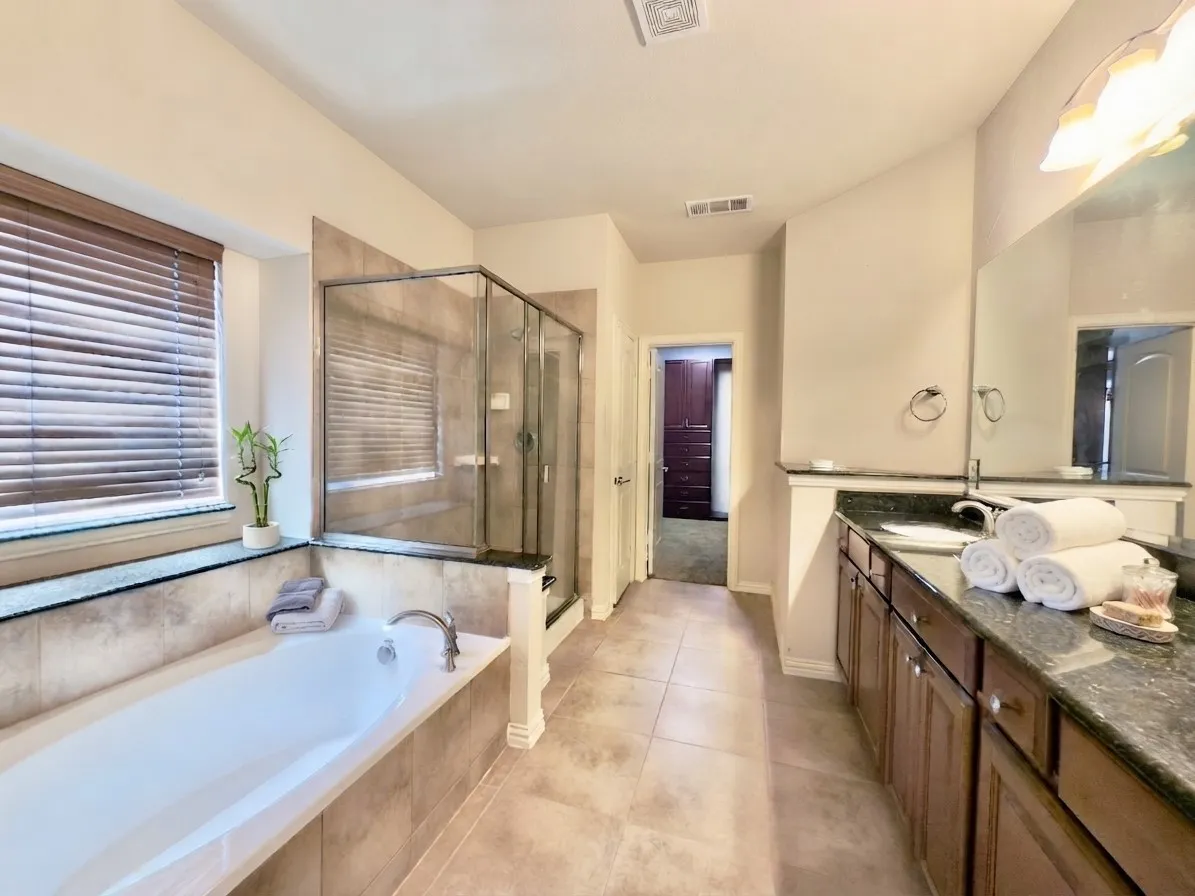 Soaking tub with a tiled surround, glass-enclosed shower, dual vanity with dark cabinetry, stone countertops, and tile flooring