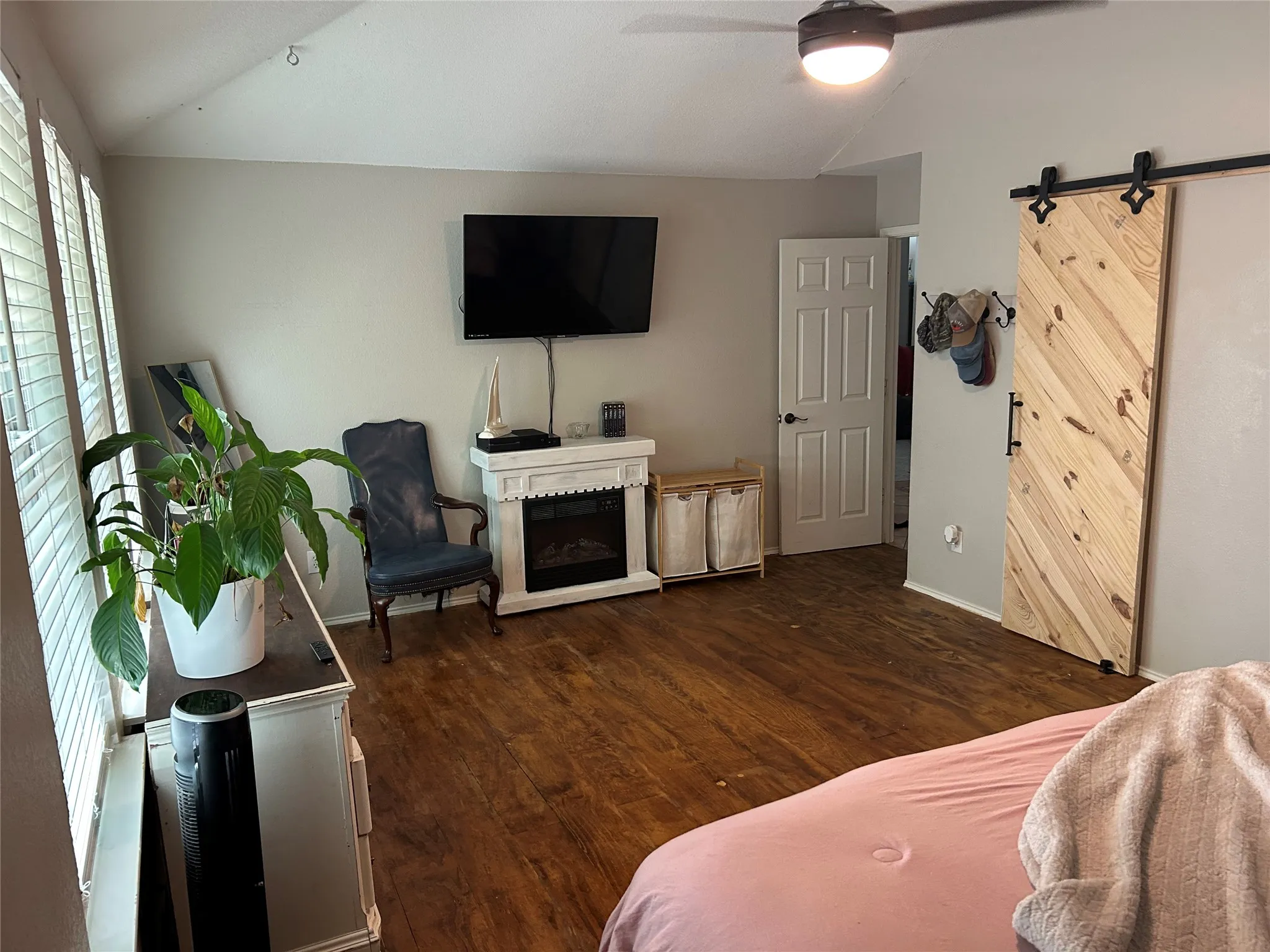 Wood-finish flooring, white fireplace mantel with electric insert, ceiling fan with integrated lighting, window with horizontal blinds, and a wood barn door on a black track