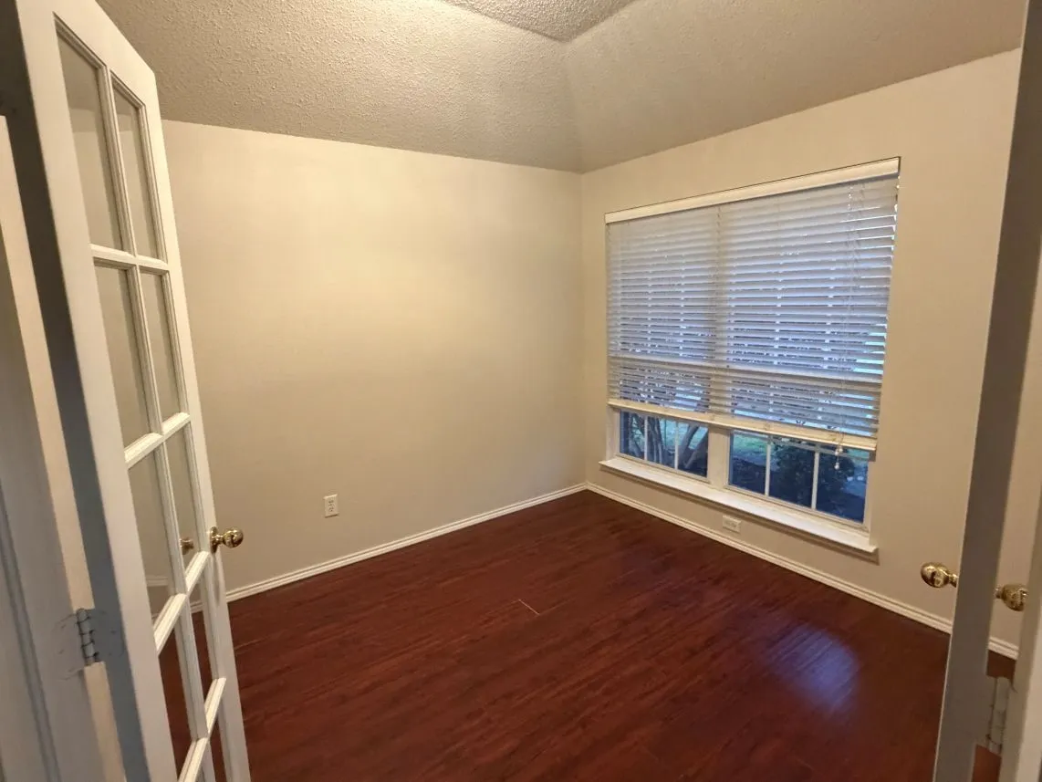 French doors with glass panels open to a room featuring wood-finish flooring, a vaulted ceiling, and a large window with blinds