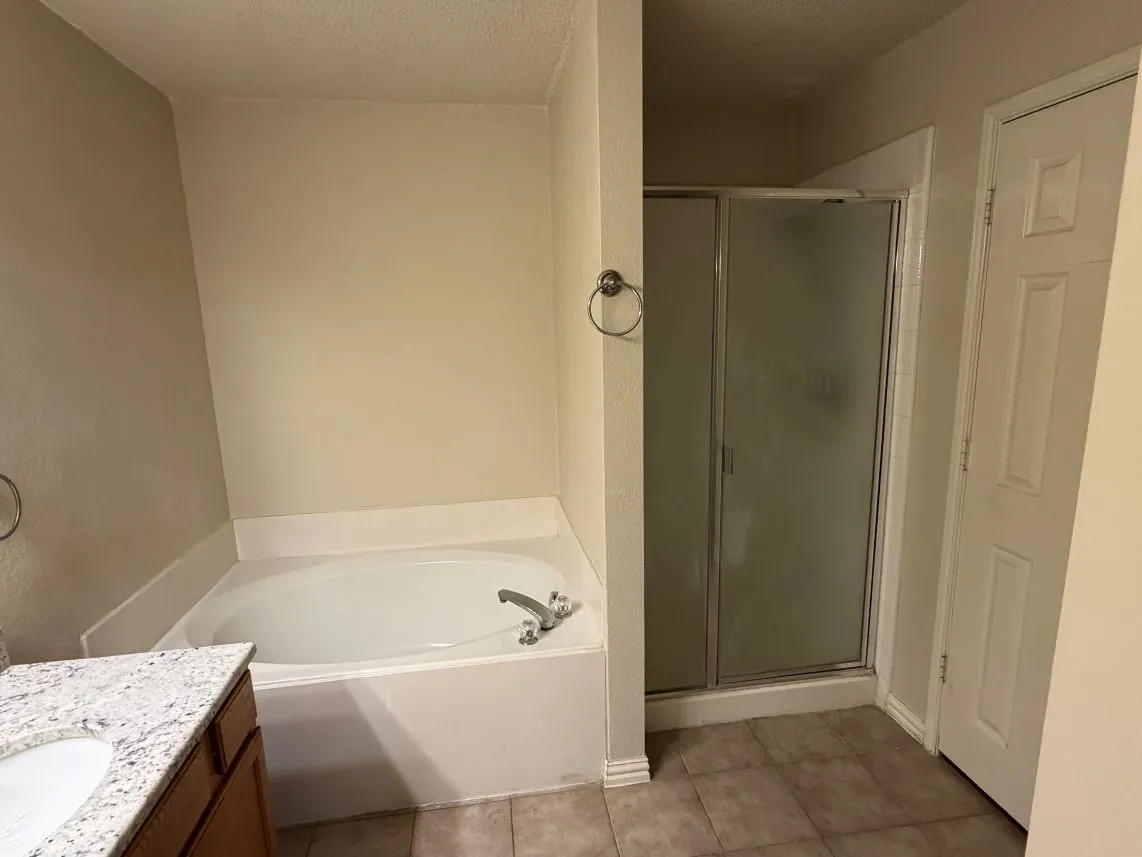 Built-in soaking tub, glass-enclosed shower, single vanity with integrated sink, stone-look countertop, and tile flooring