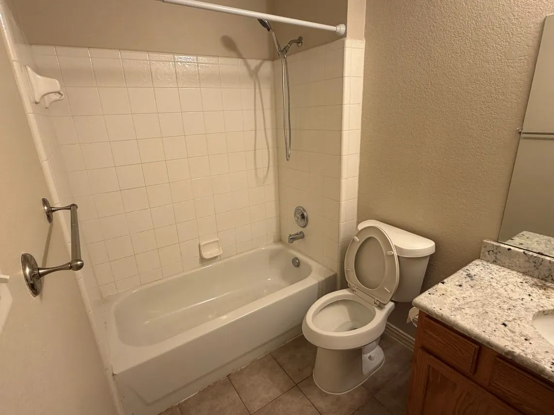 Bathtub with white tile surround, toilet, wood-finish vanity with granite-look countertop, and integrated sink
