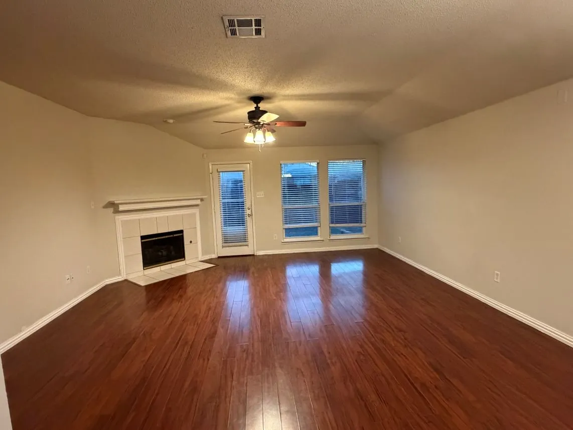 Spacious living area featuring wood-finish flooring, a tiled fireplace with mantel, multiple windows, and a ceiling fan