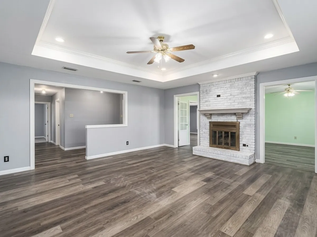 Spacious living area featuring wood-finish flooring, a white brick fireplace with a wood mantle, and a tray ceiling with recessed lighting and a ceiling fan