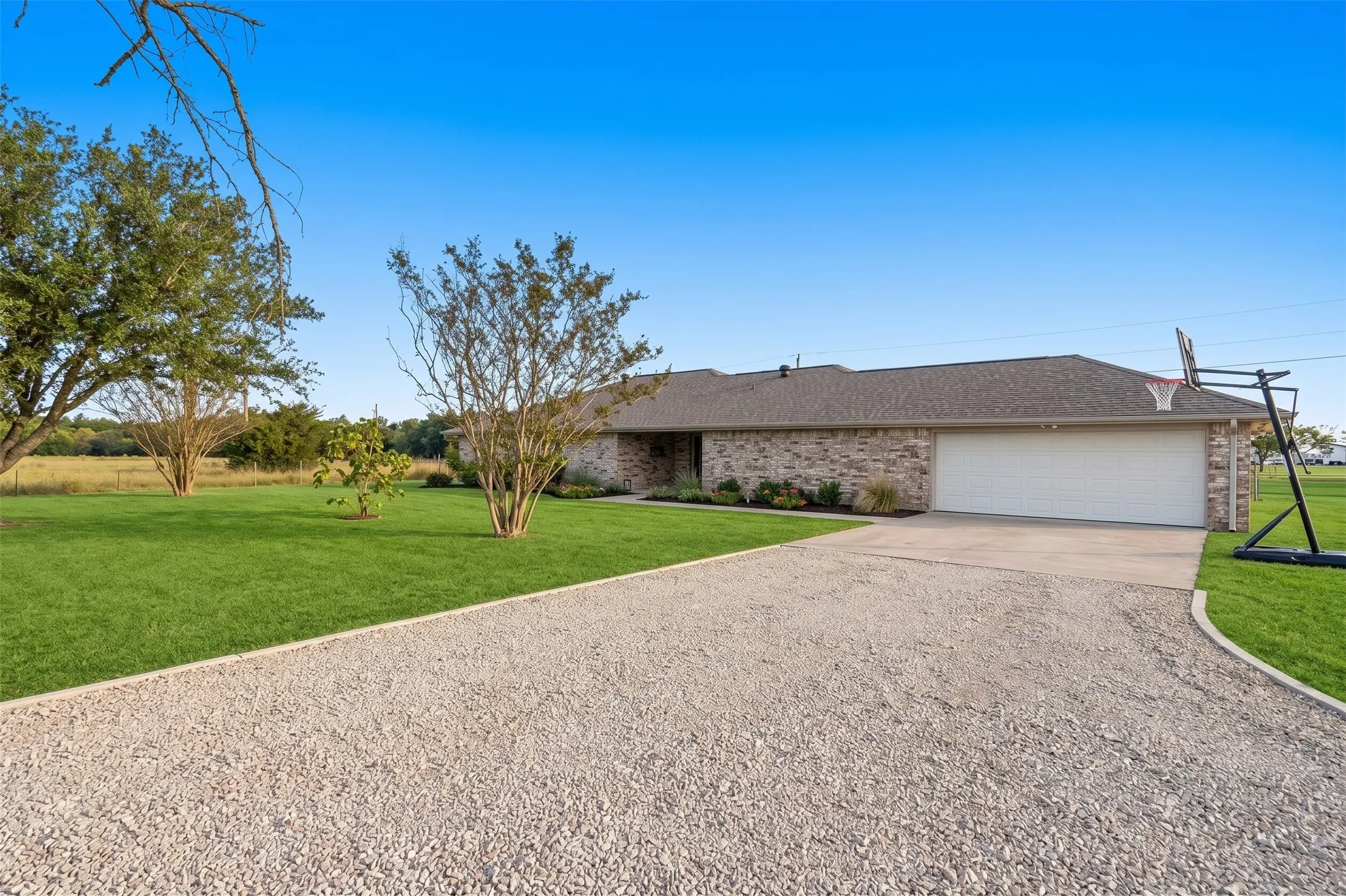 Brick exterior with a gabled roofline, attached garage, gravel driveway, and a landscaped front yard with mature trees