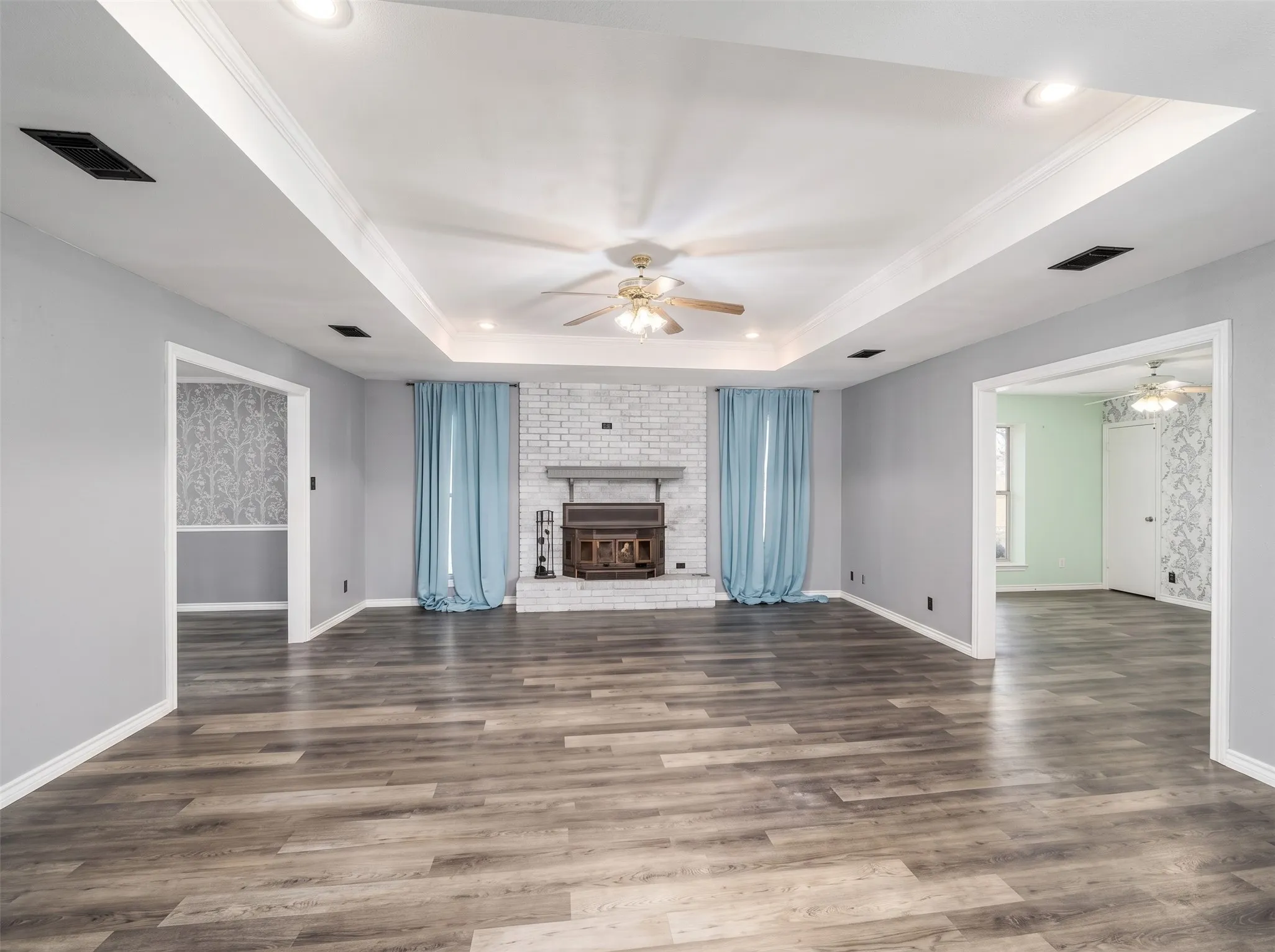 Wood-finish flooring throughout, tray ceiling with recessed lighting and ceiling fan, white brick fireplace with mantel