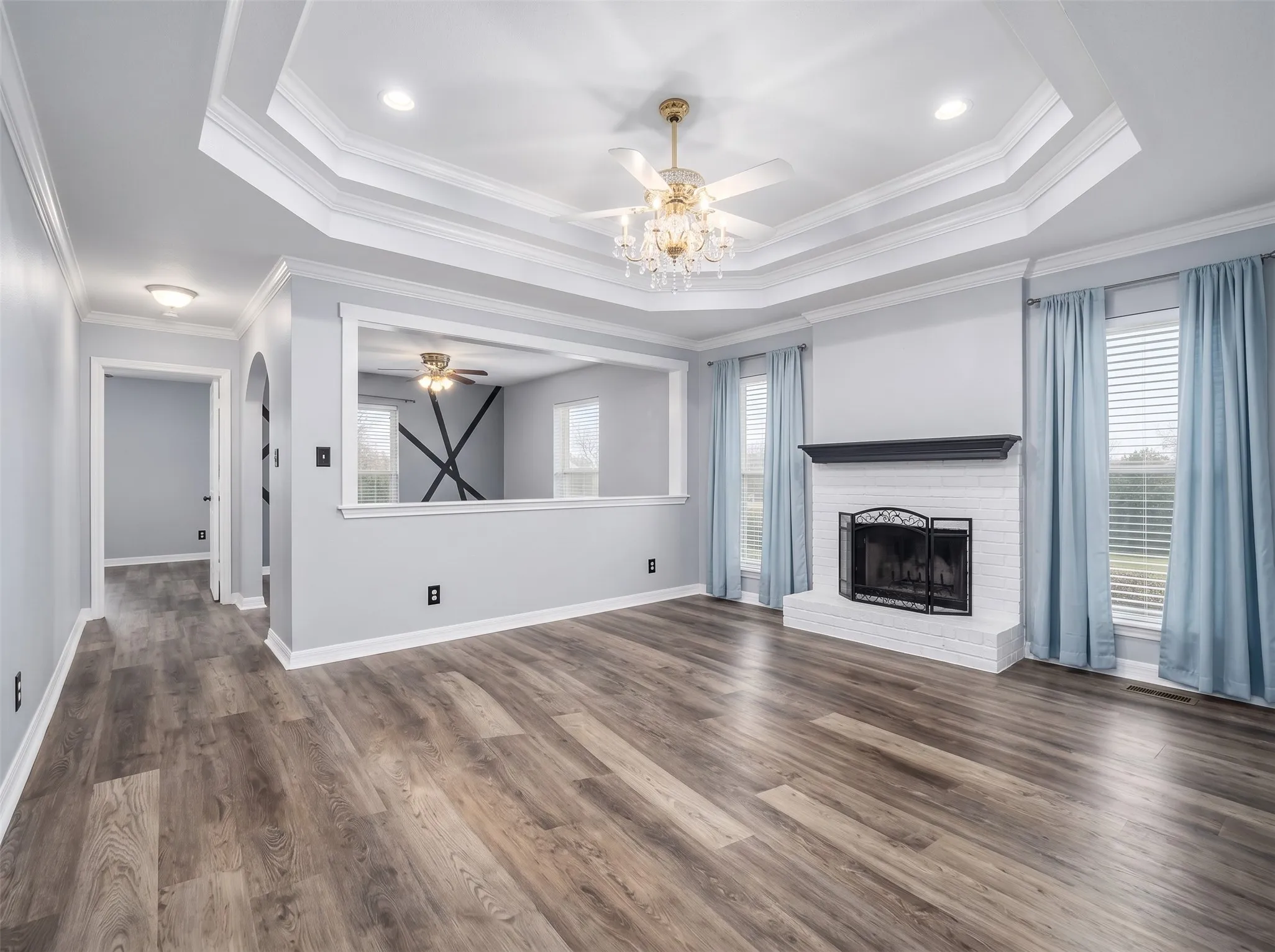 Tray ceiling with chandelier and ceiling fan, wood-finish flooring, white brick fireplace with dark mantel, arched doorway, and interior half-wall with upper opening