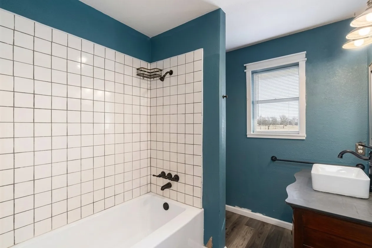 Bathtub with white subway tile surround, dark bronze shower fixtures, wood-finish flooring, and a window with blinds