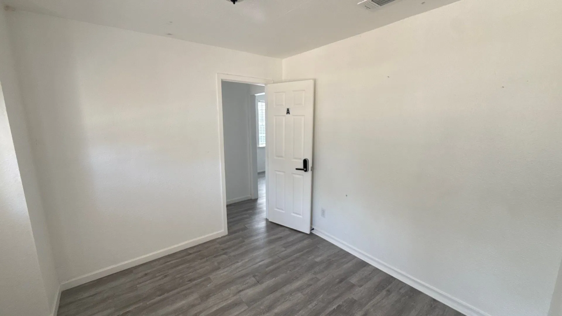 Wood-finish flooring with white baseboards, a white paneled door with black hardware, and an overhead light fixture