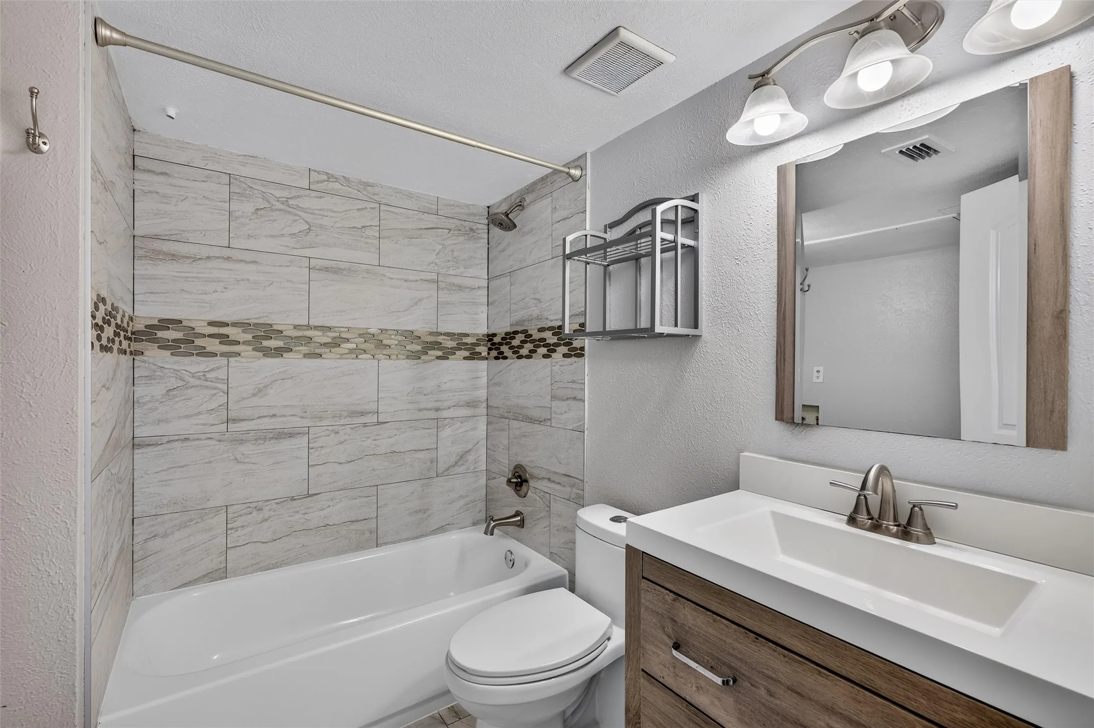 Bathroom featuring a white bathtub, gray tile shower surround with mosaic accent, wood-finish vanity with an integrated sink, and brushed nickel fixtures