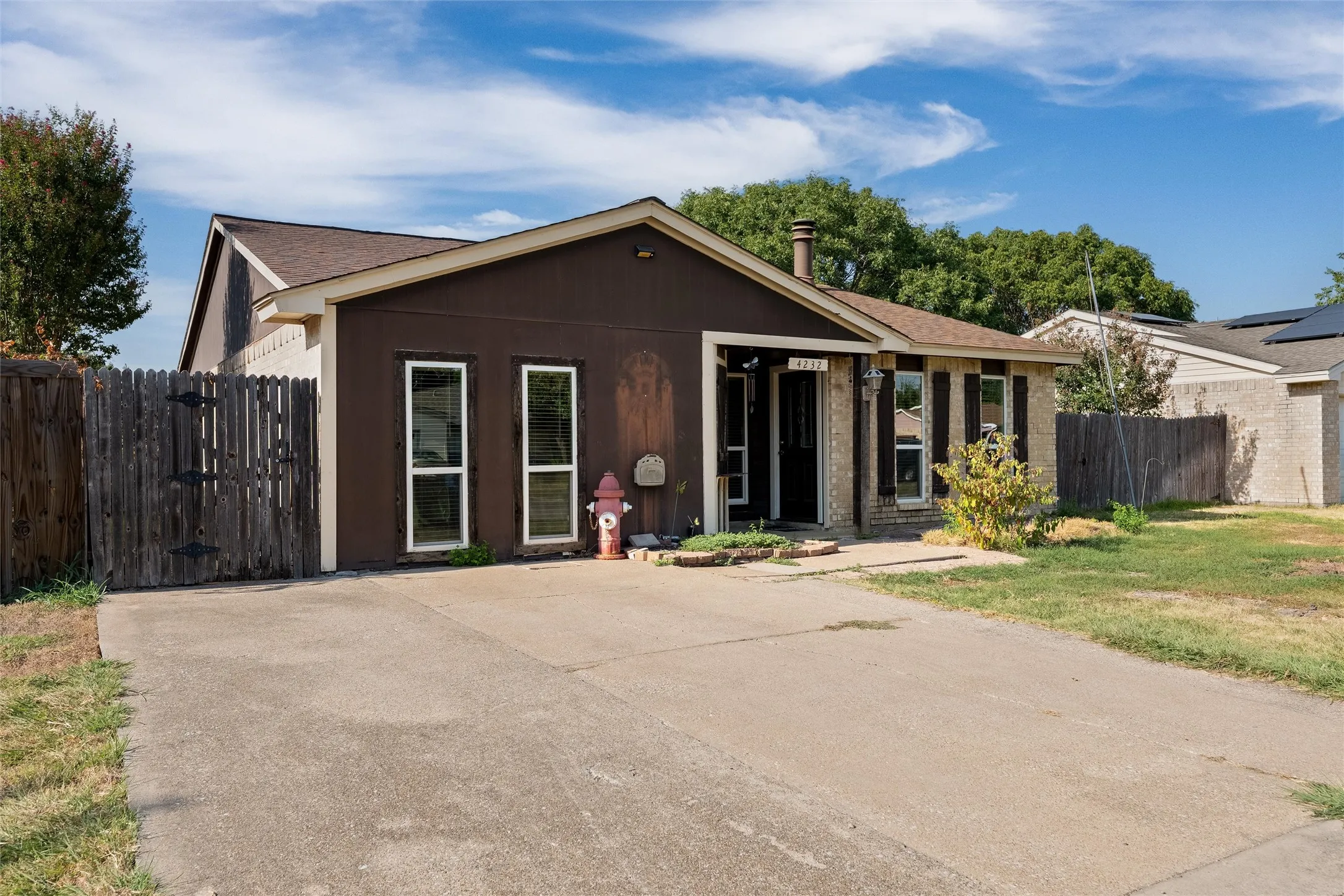 Paved driveway leading to a residence with a shingled roof, brick accents, and a wooden fence