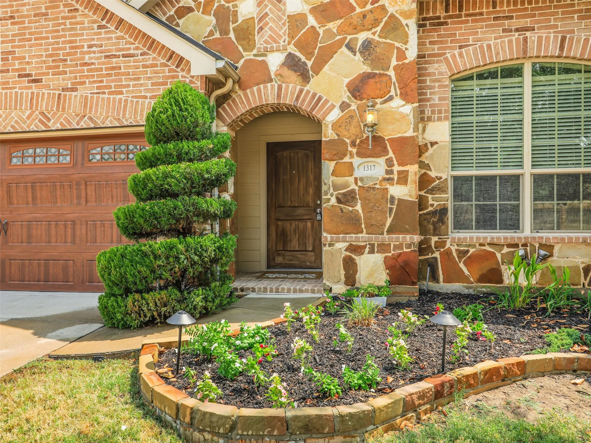A closer look at the home’s inviting covered entrance, rich stonework, decorative landscaping, and warm wood front door.
