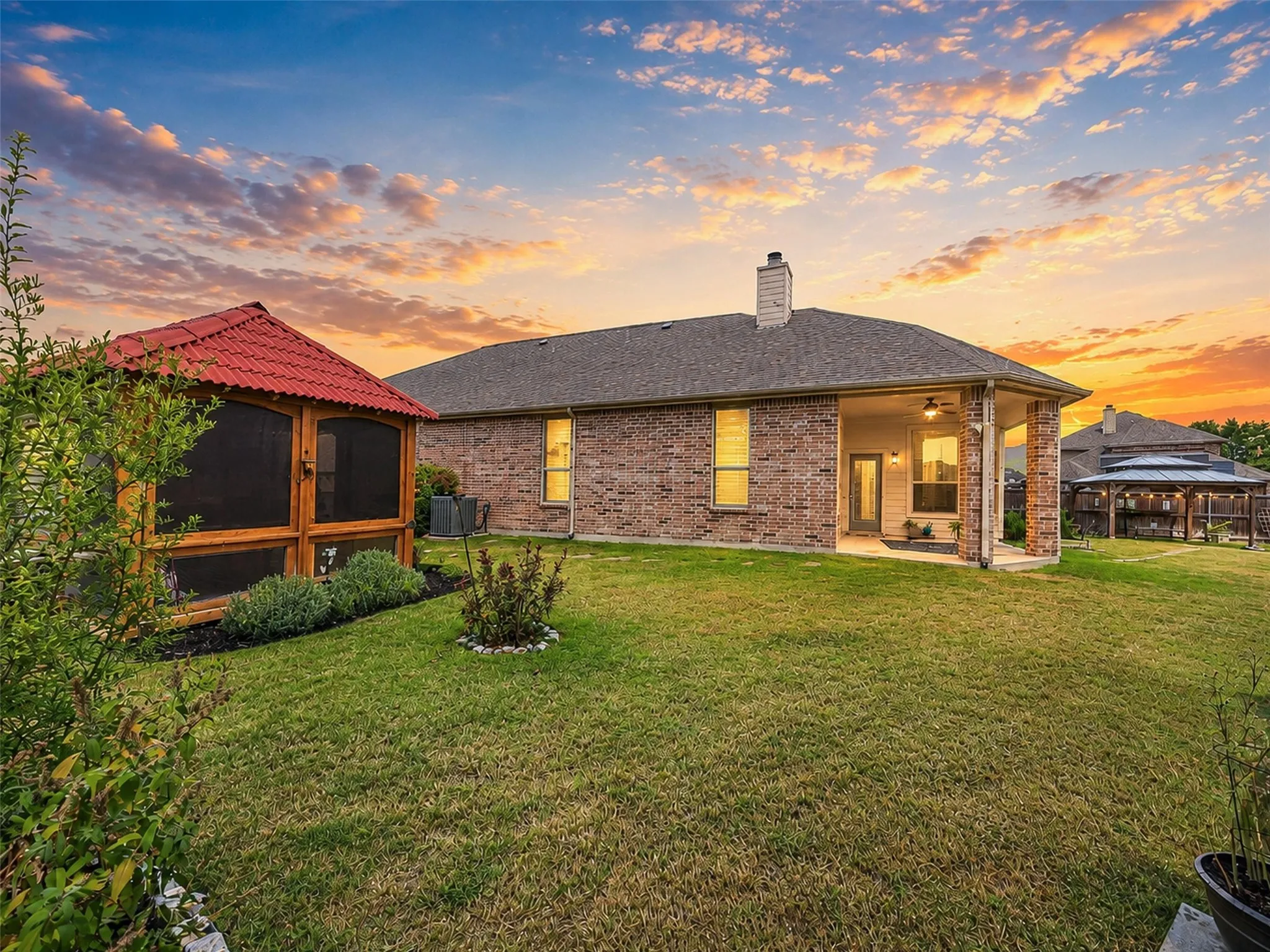 A picturesque sunset showcases the expansive backyard, covered patio, lush landscaping, gazebo, and screened outdoor retreat.