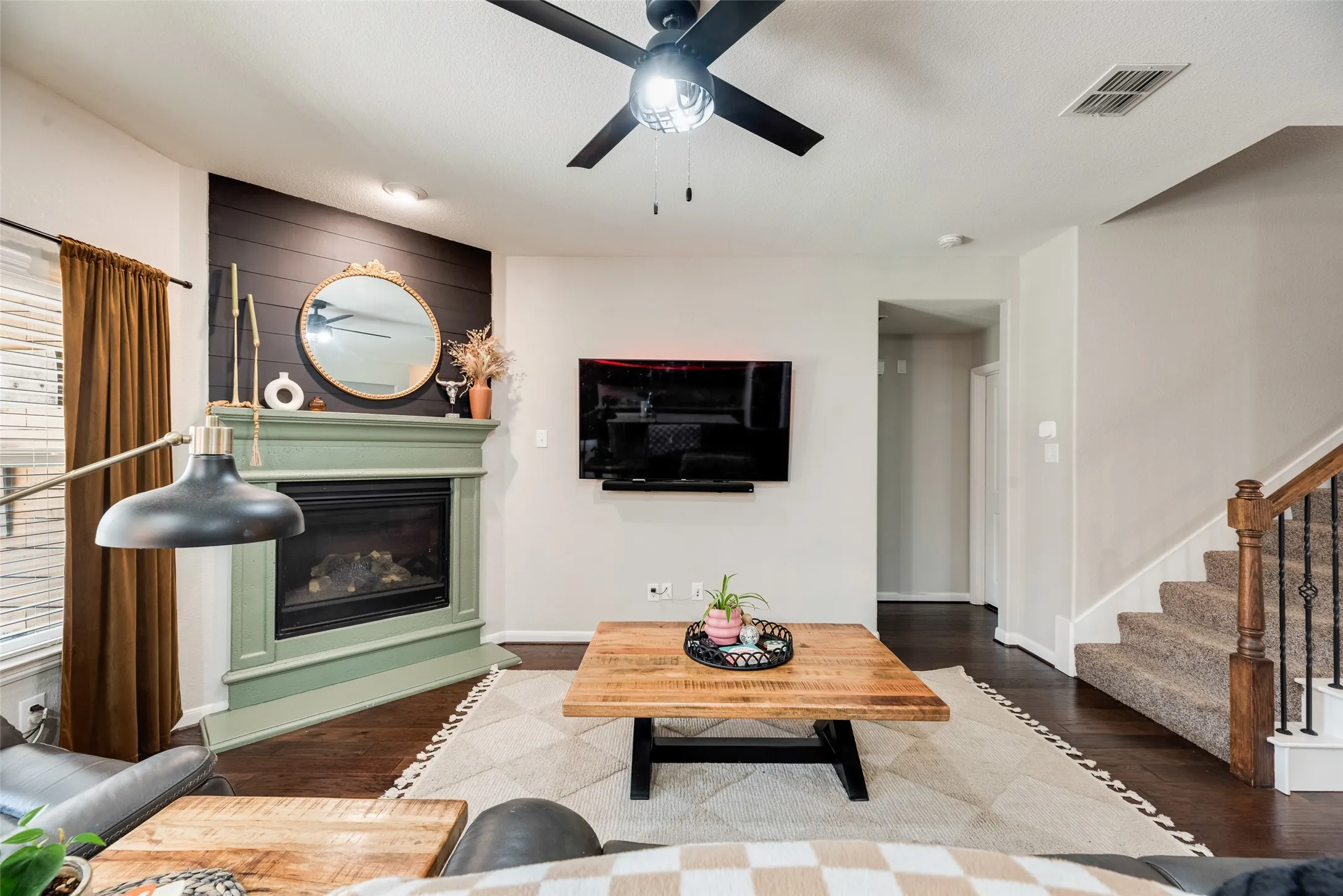 Built-in fireplace with a decorative mantel, dark wood-finish flooring, staircase with wrought iron balusters, black ceiling fan, and shiplap accent wall