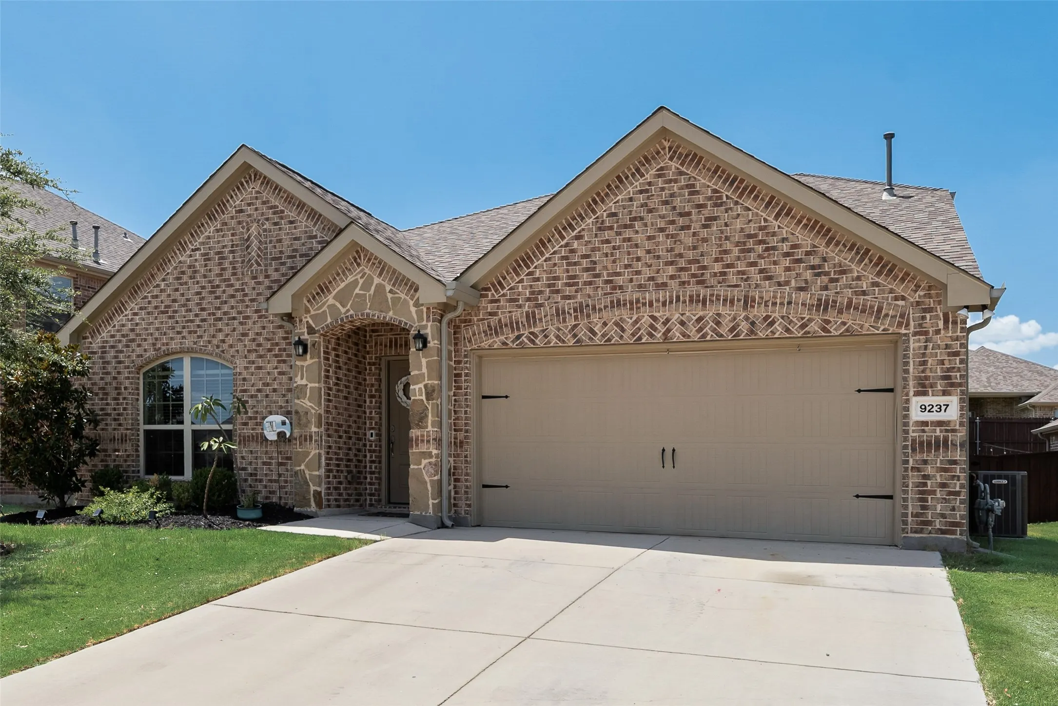 Brick and stone facade, arched entryway, attached garage, concrete driveway, and established landscaping