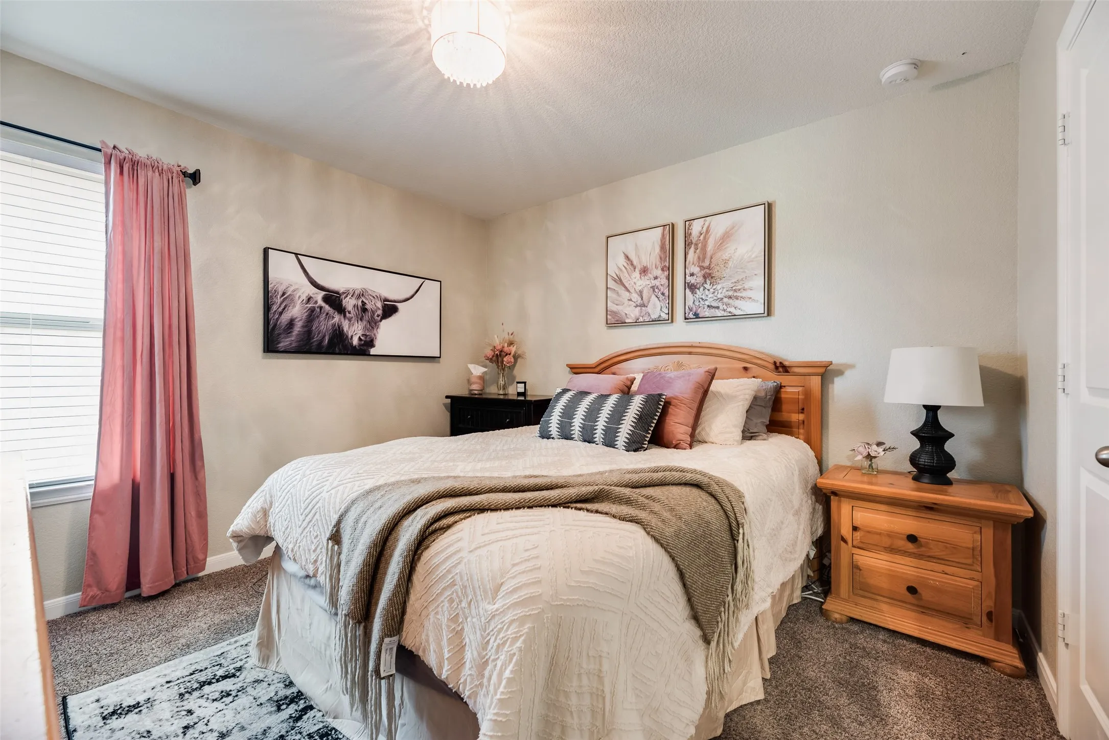 Plush carpet flooring, a natural wood headboard, a window with blinds, and an overhead light fixture