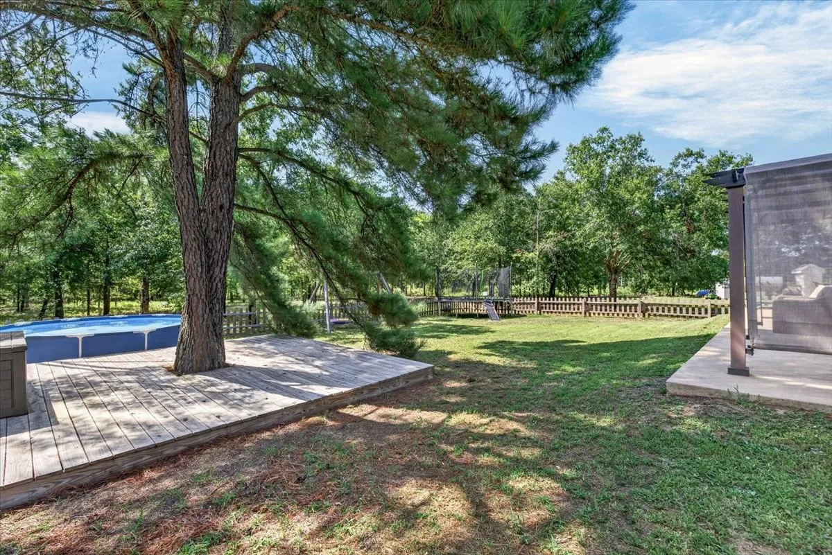 Expansive backyard featuring a large tree, above-ground pool with surrounding wood decking, concrete patio, and fenced perimeter