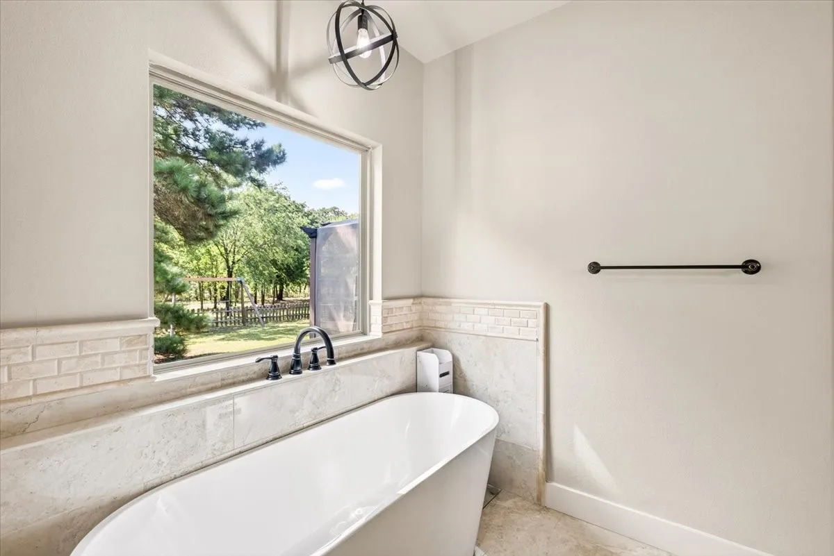 Freestanding soaking tub with matte black fixtures, framed window, beige subway tile detailing, and an industrial-style overhead light fixture