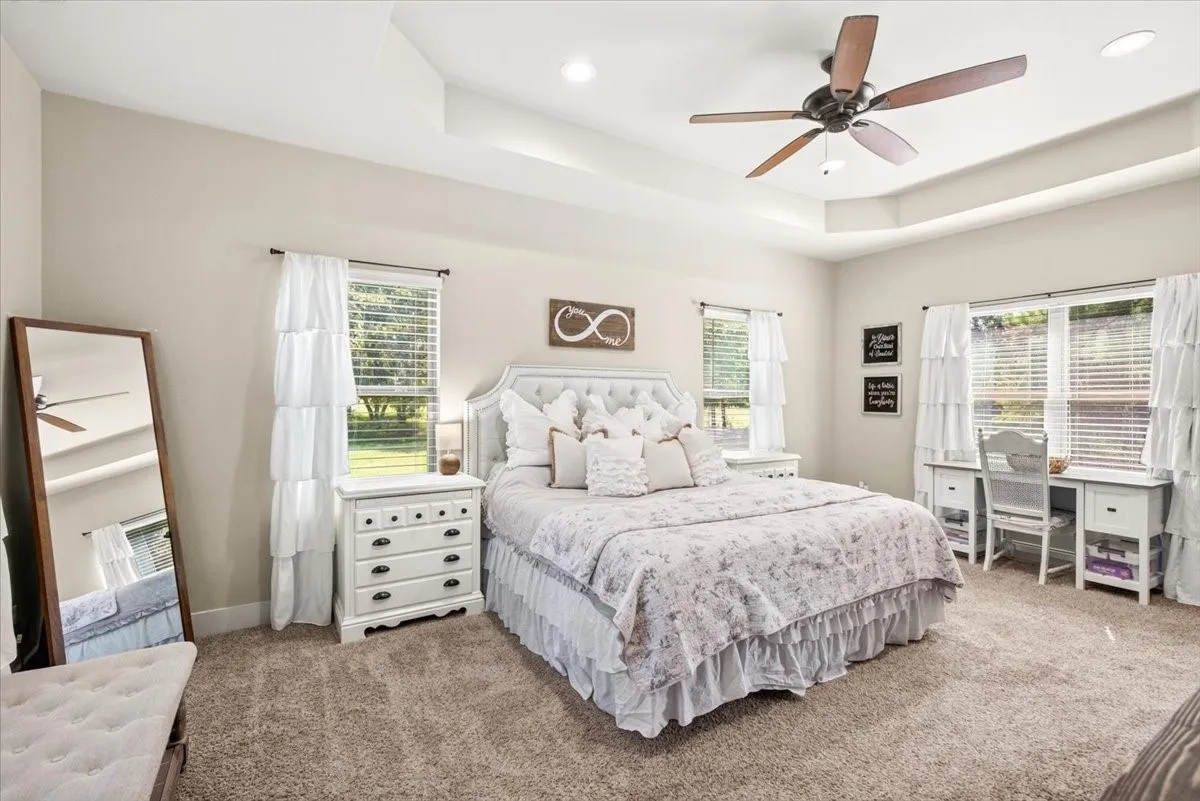 Tray ceiling with recessed lighting, ceiling fan, tufted headboard, double windows with blinds, and neutral carpeting