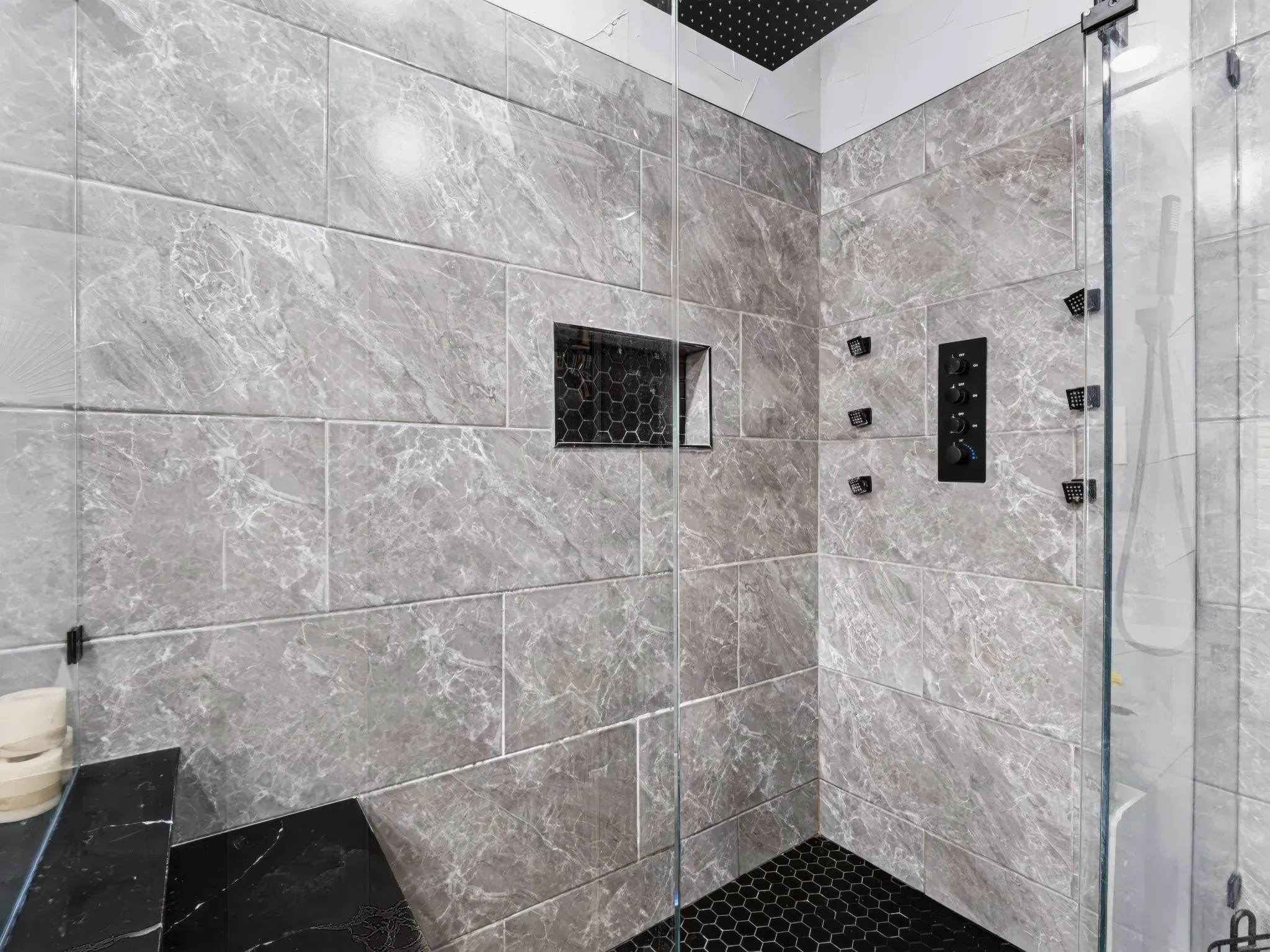 Glass-enclosed shower featuring large format wall tiles, a built-in shower niche with hex tile, a rainfall showerhead, and multiple body sprayers