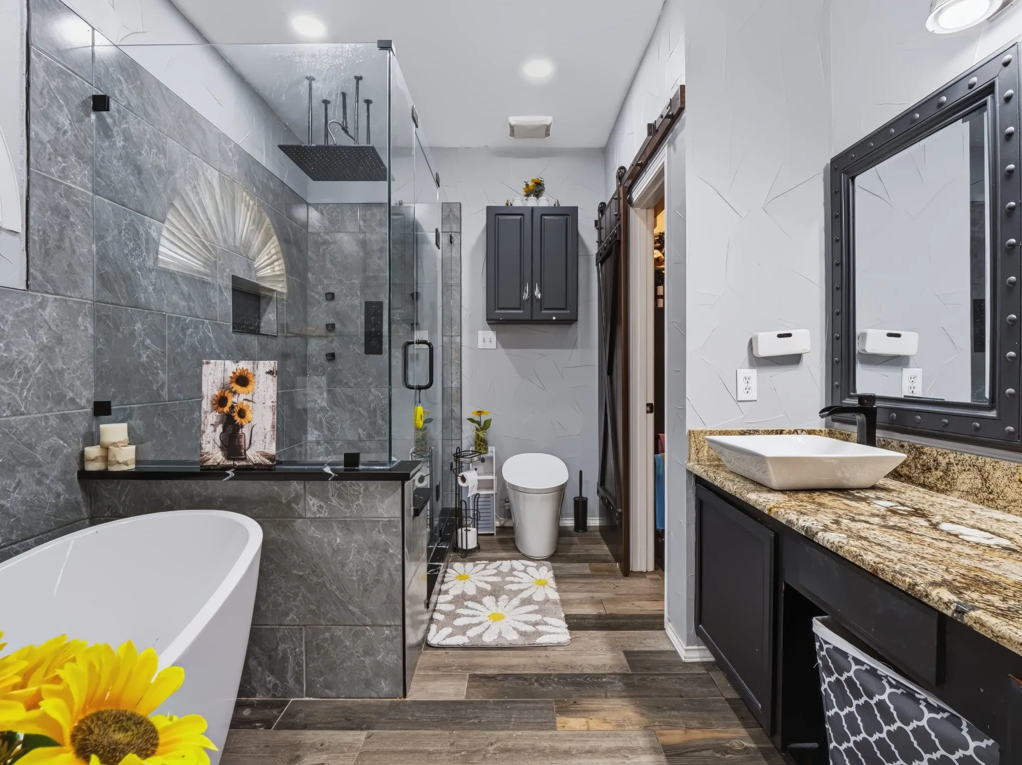 Glass-enclosed shower with a rainfall showerhead, freestanding soaking tub, wood-finish flooring, vanity with a vessel sink, and a barn door entry